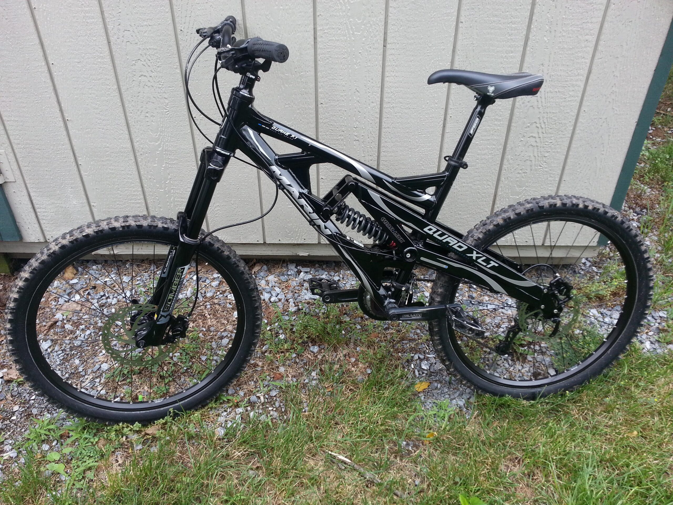 Marin 2007 Marin Quake 7.1: A black and white mountain bike with a full suspension system, set against a textured wooden background. The bike features knobby tires, disc brakes, and a sleek frame design, placed on a grassy area with small stones visible around the base.