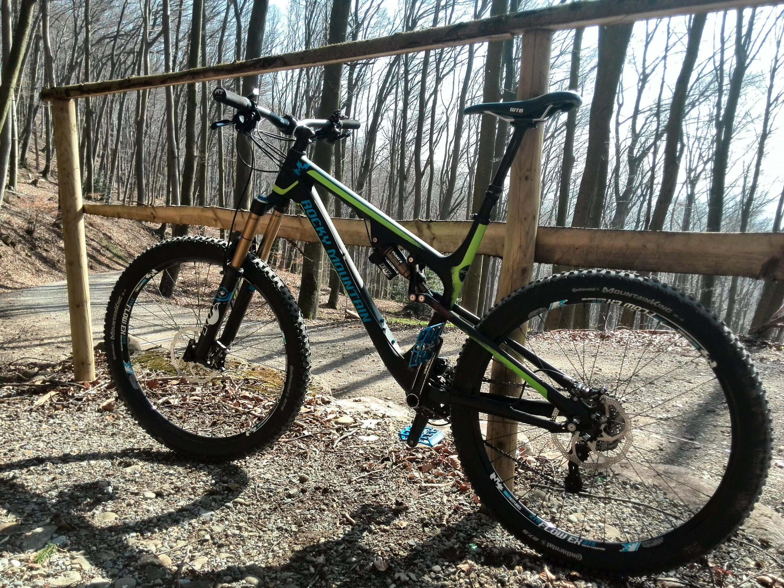 Rocky Mountain Altitude 750: A mountain bike leaning against a wooden fence on a gravel path, surrounded by tall trees in a forest setting. The bike features thick tires, a black and green frame, and is positioned in sunlight filtering through the trees.