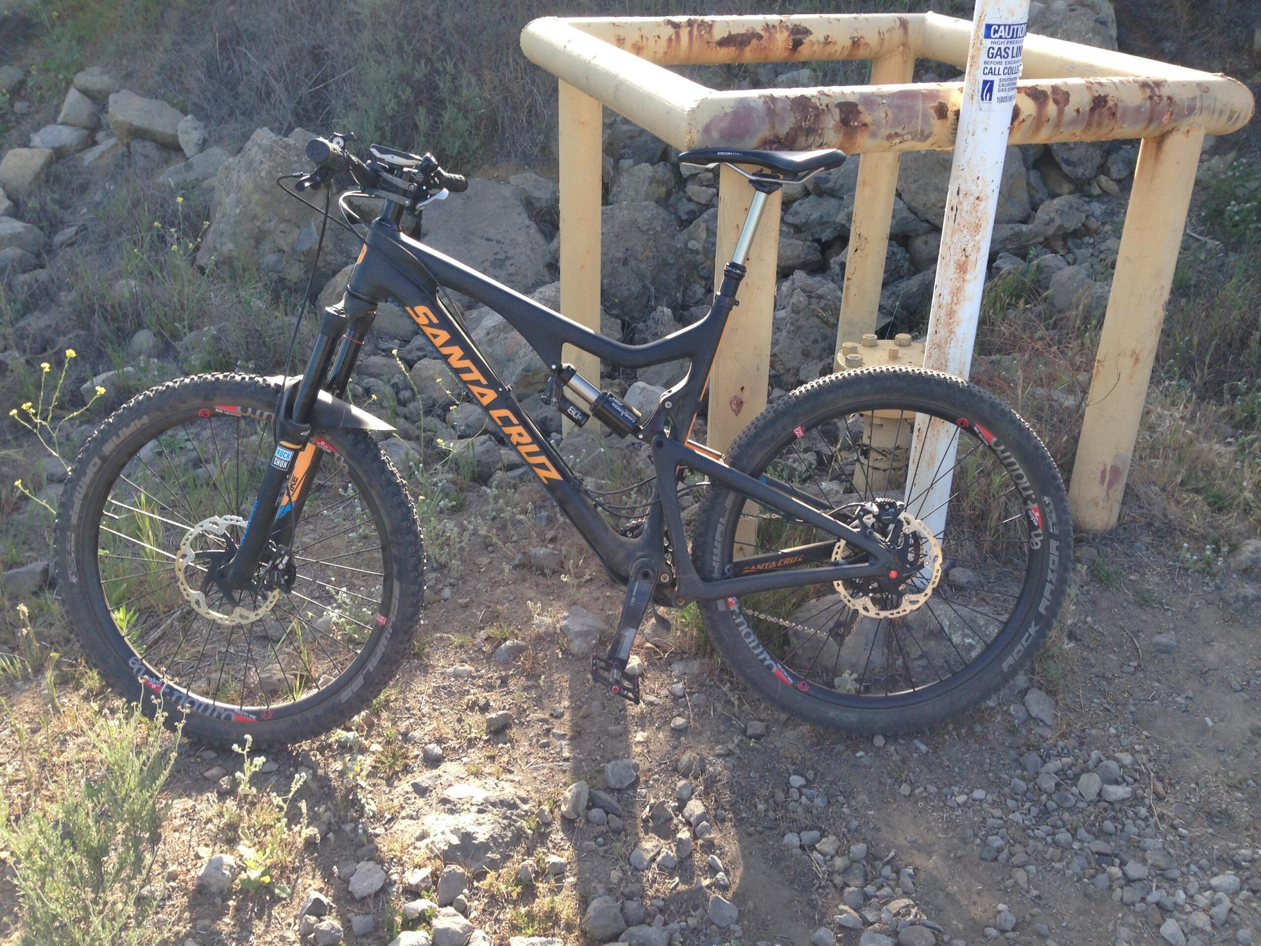 Santa Cruz Bronson C: A black Santa Cruz mountain bike resting on rocky terrain, with a yellow safety structure visible in the background. The bike features thick tires and prominent branding, surrounded by grass and small plants.