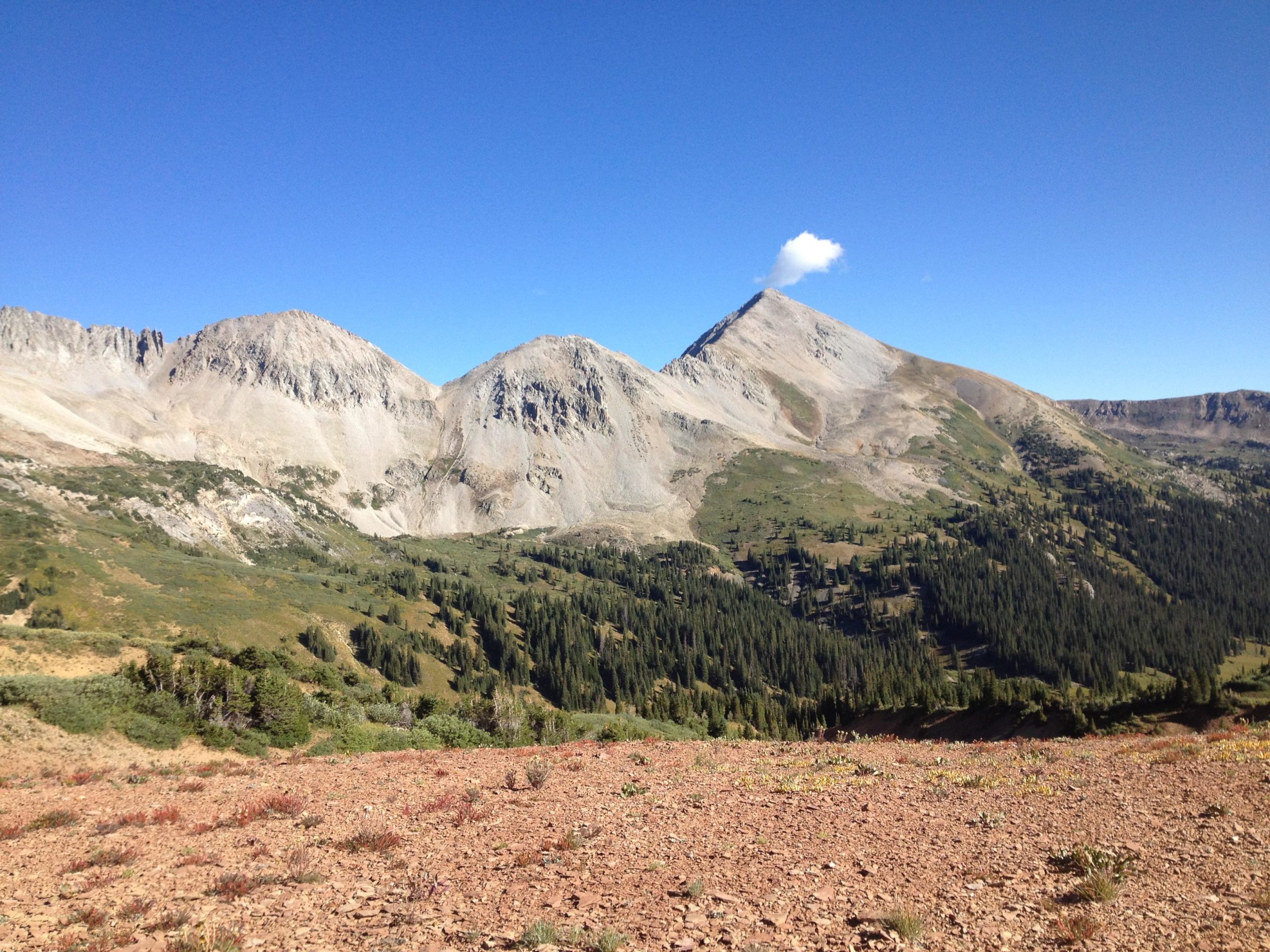 A scenic view of a mountainous landscape featuring multiple peaks under a clear blue sky. The foreground has rocky terrain with patches of vegetation, while the background showcases steep, rugged mountains with a small cloud perched on the peak of one mountain. Lush green forests are visible in the valleys between the mountains. Crystal Peak mountain bike trail.
