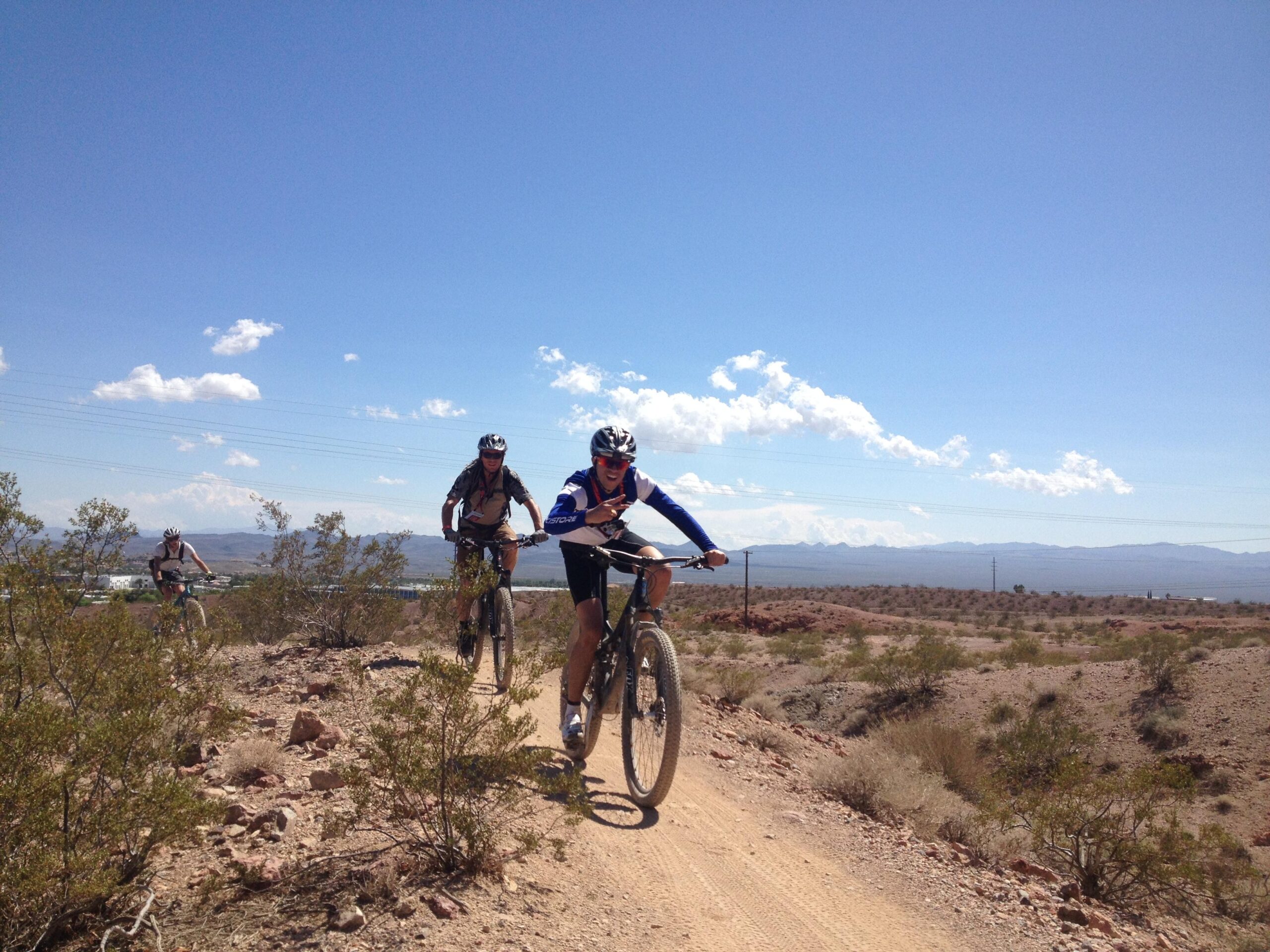 Three mountain bikers ride along a dirt trail in a desert landscape, with sparse vegetation and hills in the background. The sky is bright blue with a few clouds. Bootleg Canyon mountain bike trail.