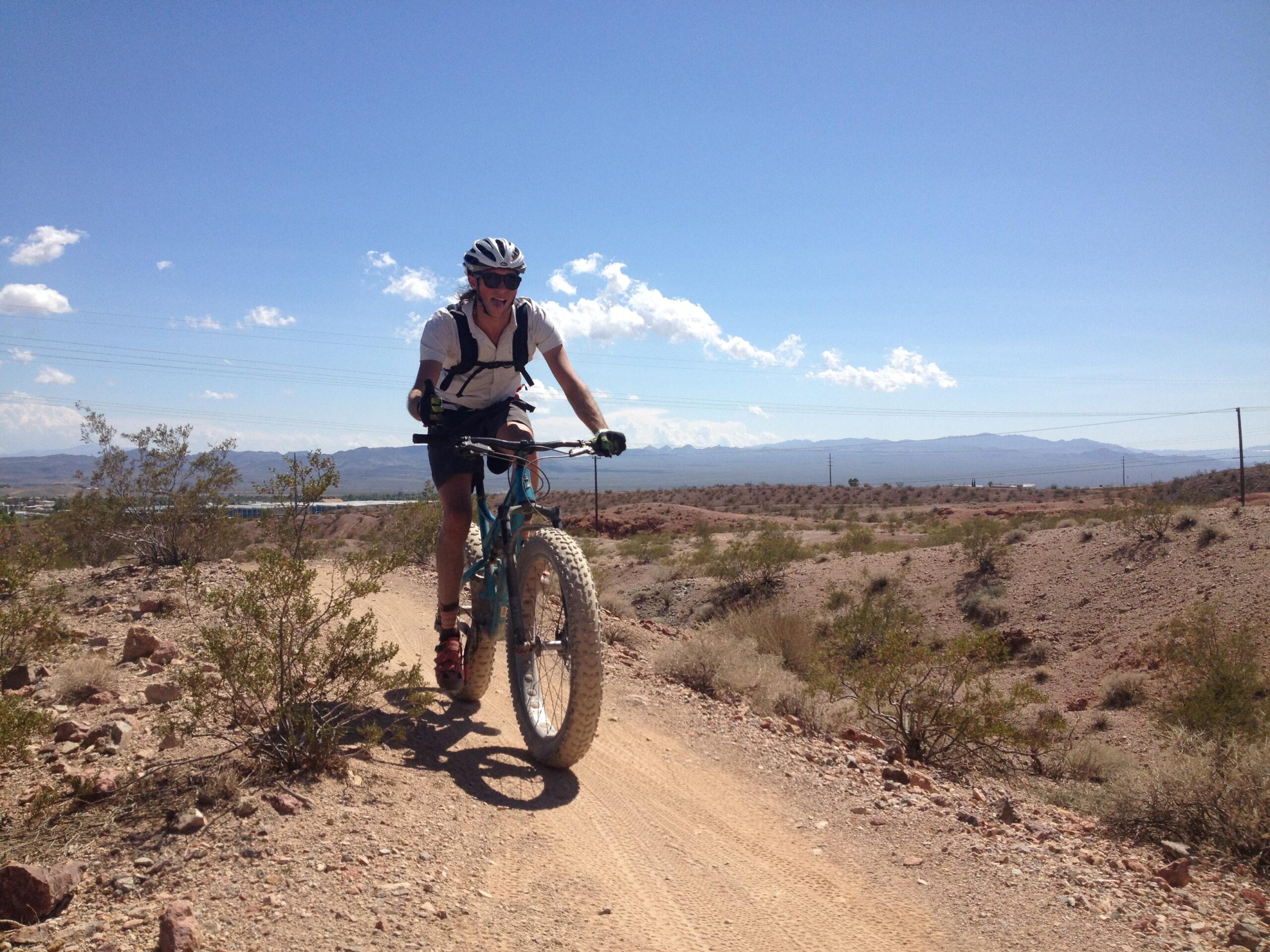 A cyclist riding a turquoise fat bike on a dirt trail in a desert landscape, with mountains visible in the background and a clear blue sky above. The cyclist is wearing a helmet, sunglasses, and a backpack, and appears to be enjoying the ride. Surrounding vegetation includes sparse bushes and rocky terrain. Bootleg Canyon mountain bike trail.