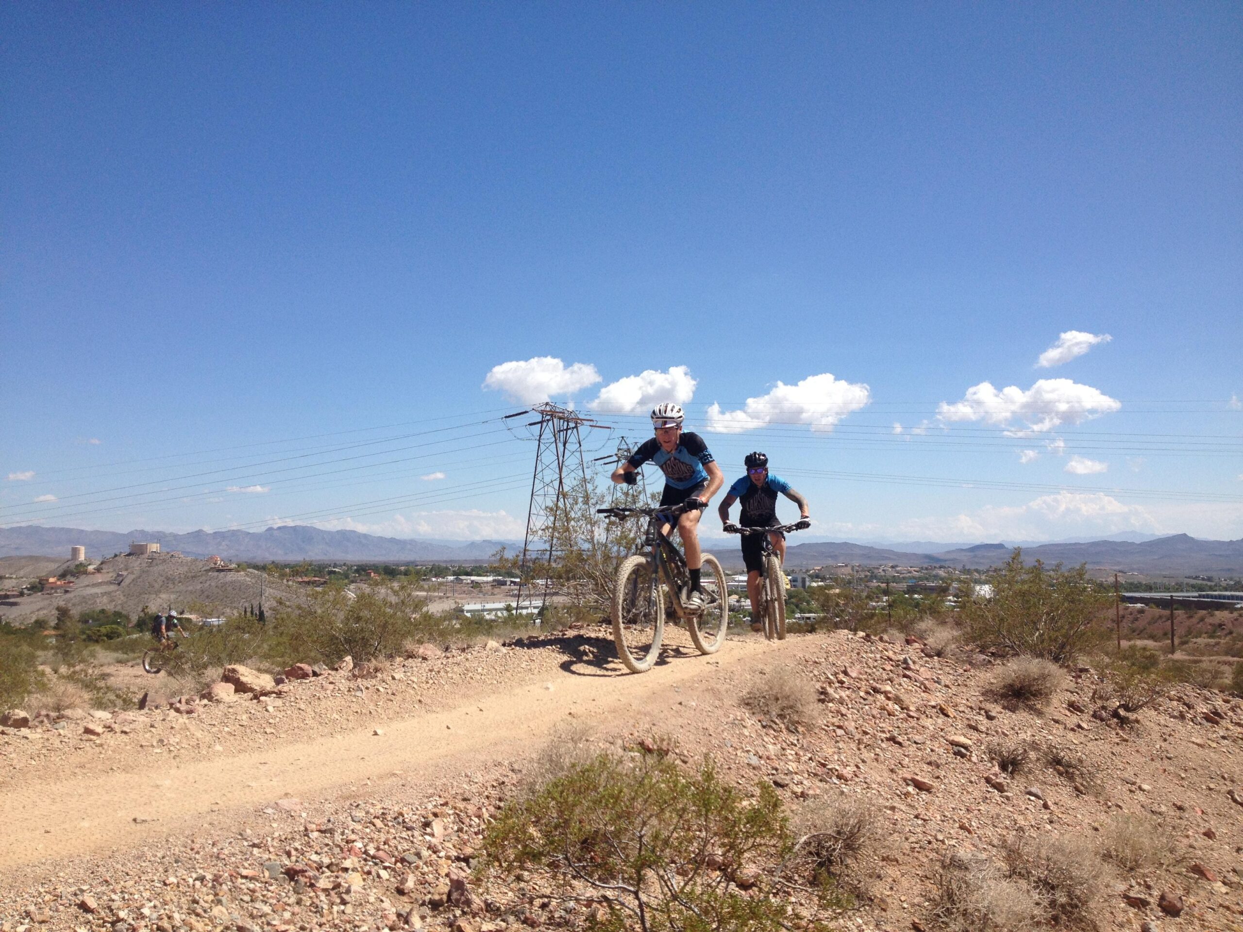 Two mountain bikers navigate a dirt trail in a rugged landscape under a clear blue sky. Power lines run in the background, and distant mountains are visible. The terrain features sparse vegetation and rocky ground. Bootleg Canyon mountain bike trail.