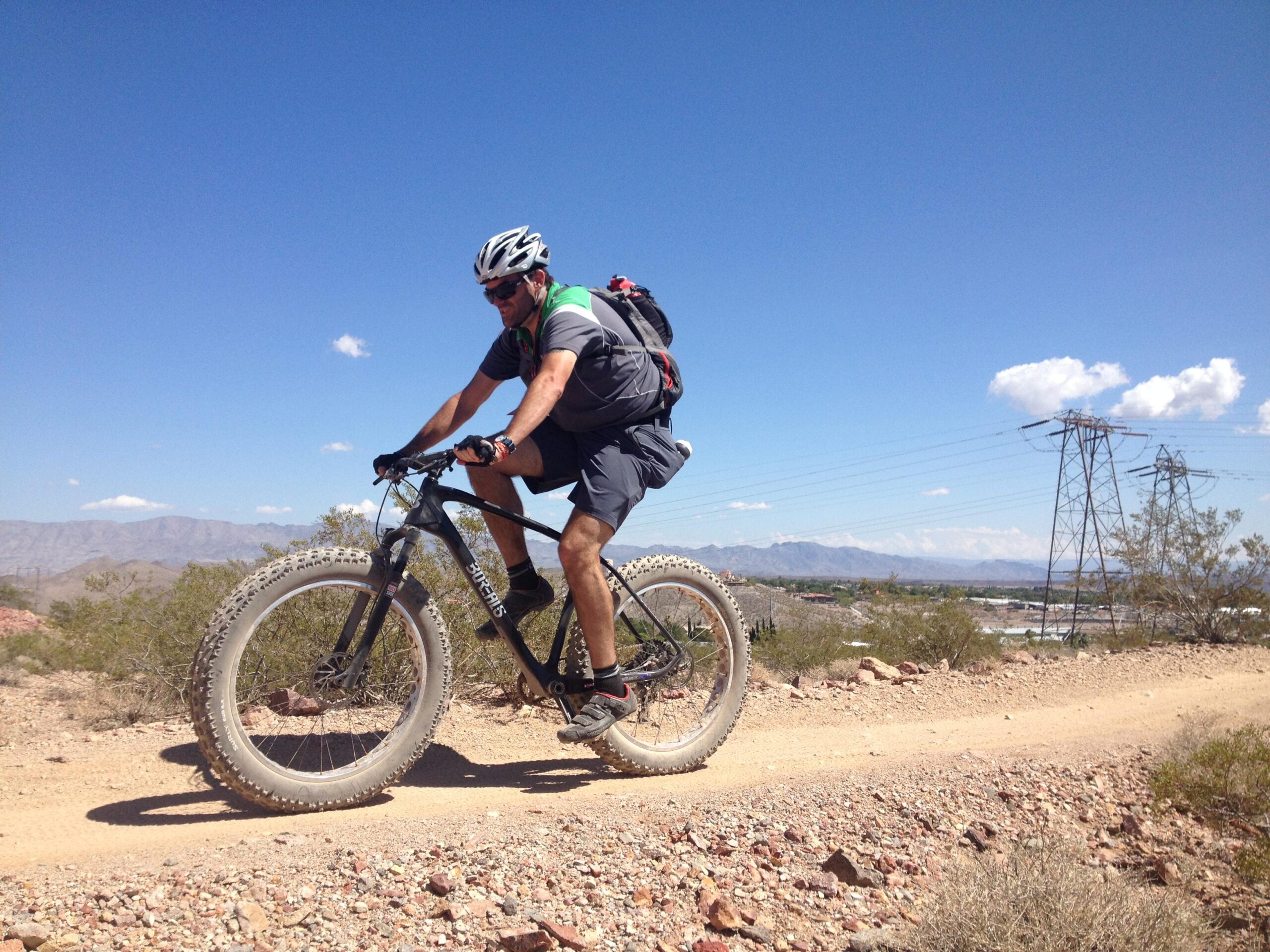 A cyclist riding a fat bike on a rocky dirt trail under a clear blue sky. The landscape features sparse desert vegetation and mountains in the background, with power lines visible in the distance. The rider is wearing a helmet and a backpack, dressed in athletic clothing. Bootleg Canyon mountain bike trail.
