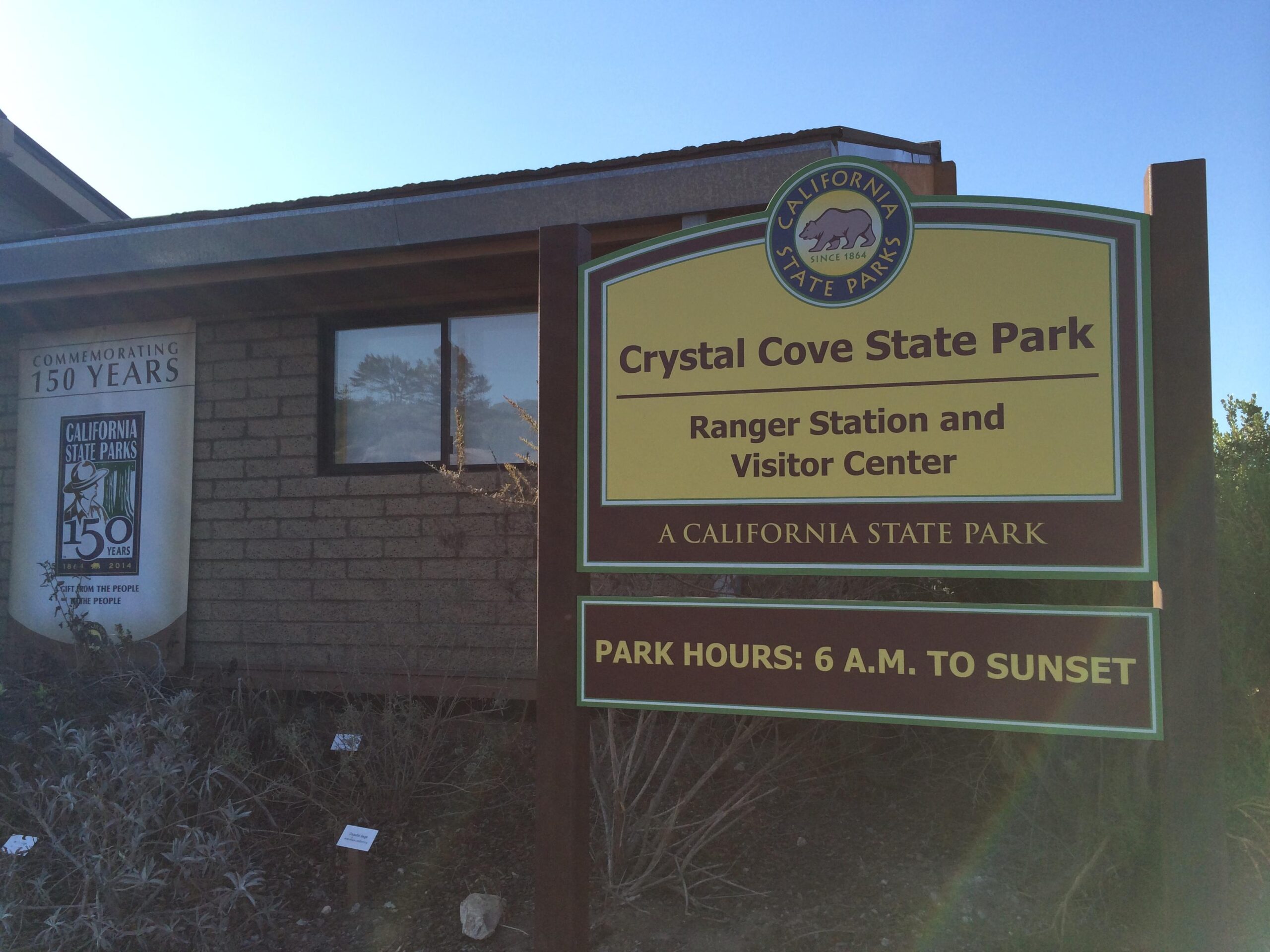 Signage for Crystal Cove State Park, indicating the Ranger Station and Visitor Center. The sign features a bear emblem and highlights park hours from 6 A.M. to sunset. A banner commemorates the 150th anniversary of California State Parks. Crystal Cove State Park mountain bike trail.