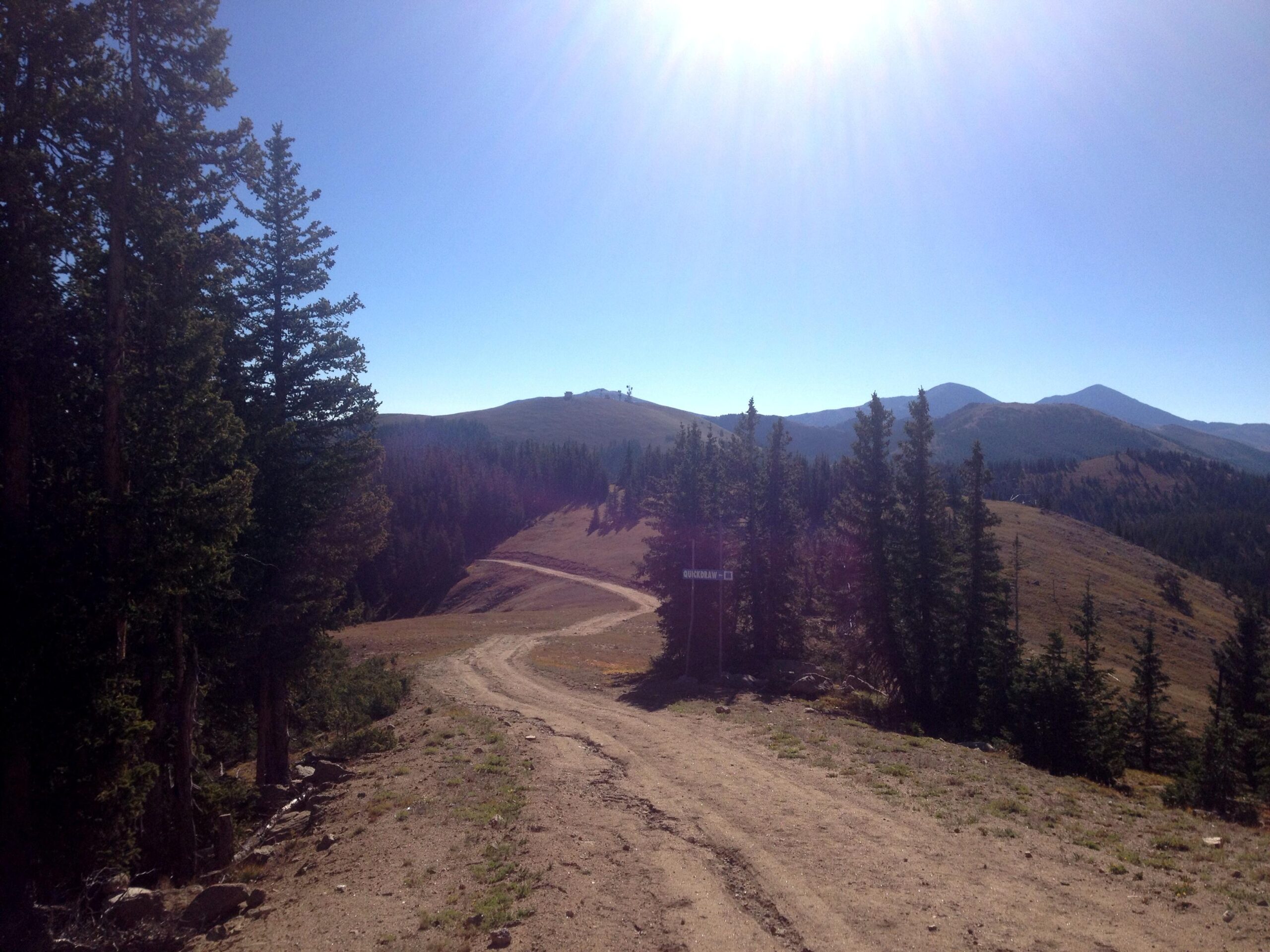 A sunlit dirt road winding through a mountainous landscape, bordered by tall evergreen trees on both sides. The sky is clear blue with the sun shining brightly, and distant hills are visible in the background. A sign indicating a trail name is partially visible along the road. CDT: Monarch Pass to Boss Lake / Hunt Lake Trail mountain bike trail.