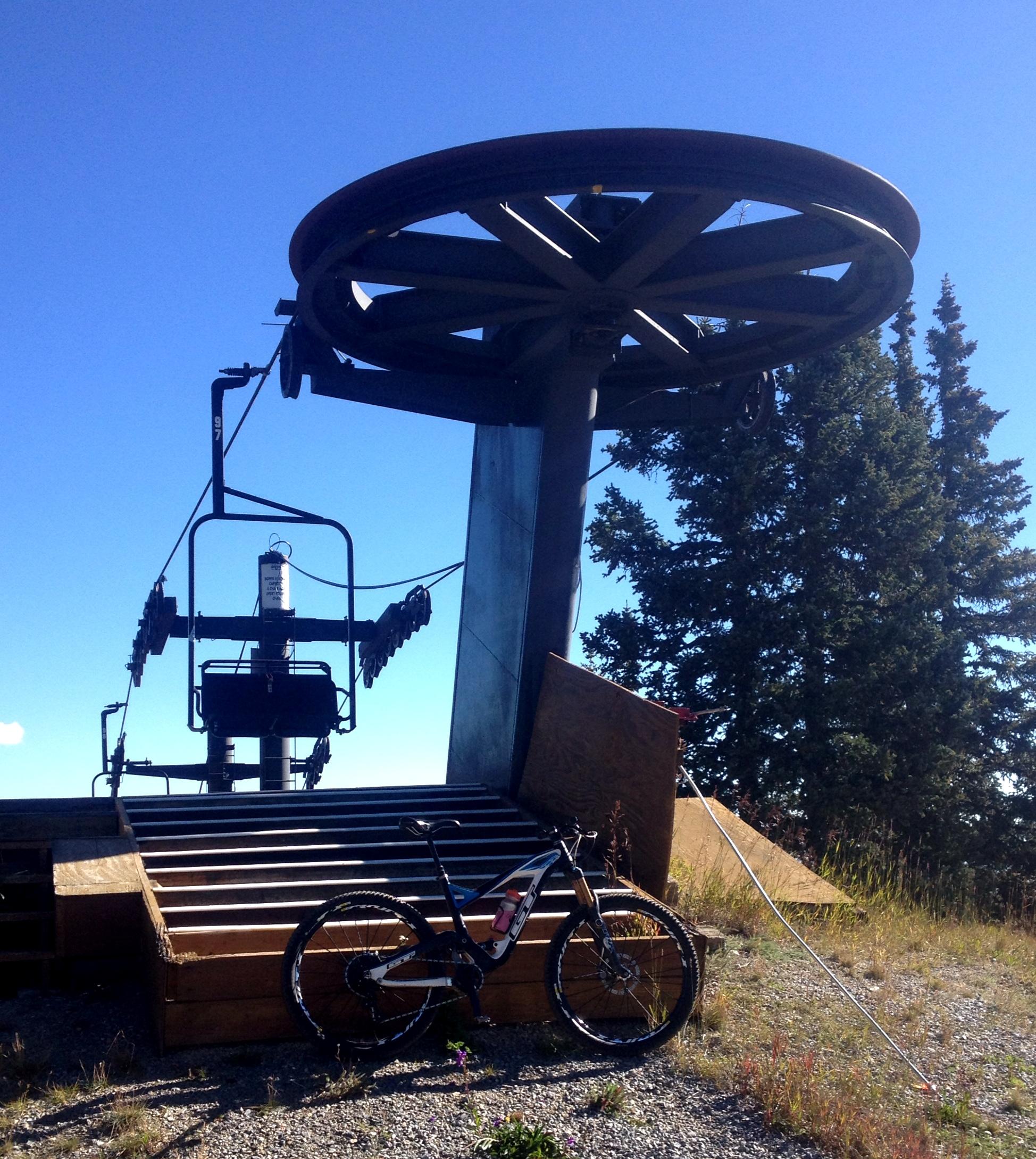 A mountain bike resting on gravel near the base of a ski lift, featuring a large circular wheel and metal supports against a clear blue sky and evergreen trees in the background. CDT: Monarch Pass to Boss Lake / Hunt Lake Trail mountain bike trail.