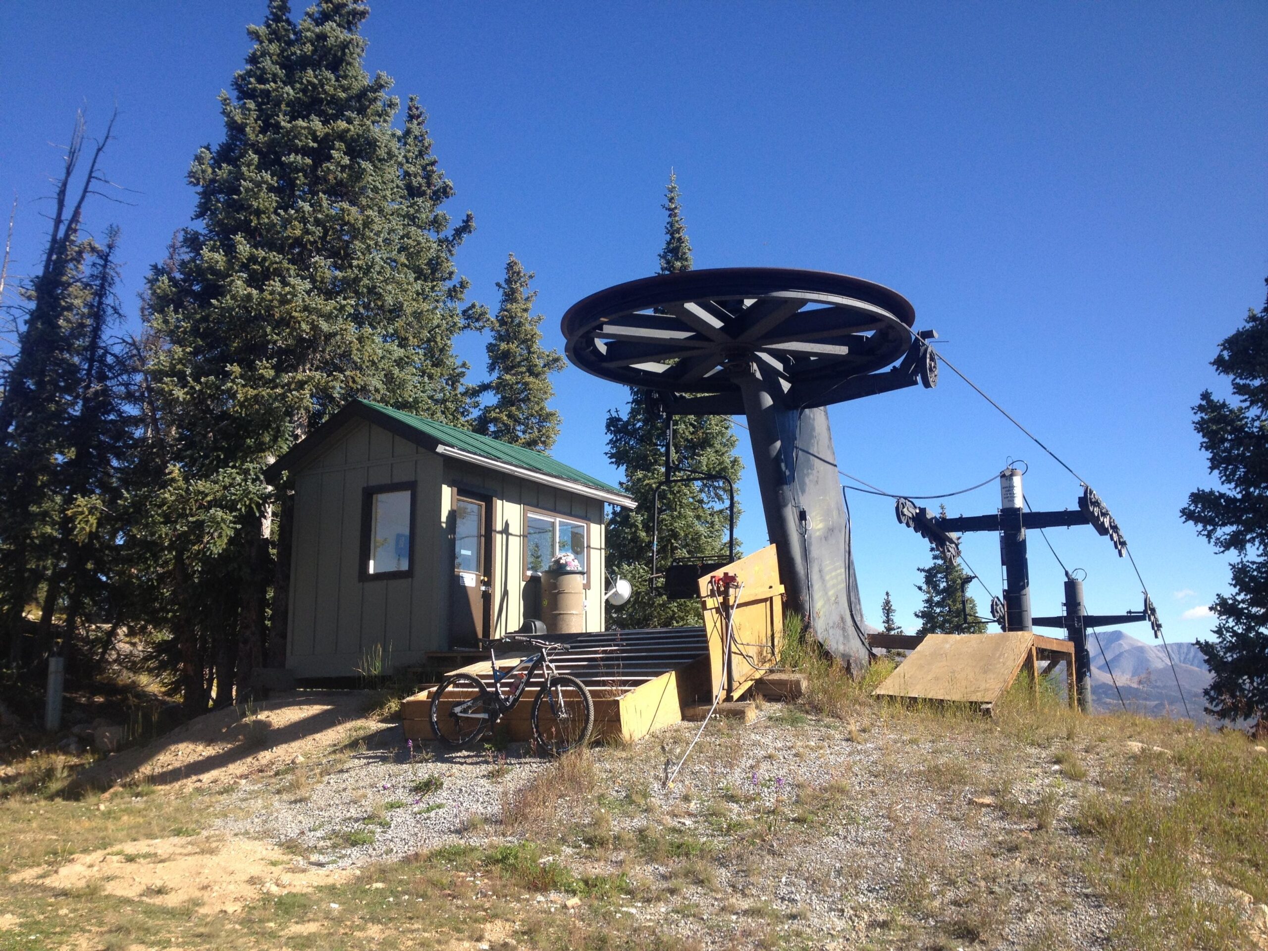 Cable car station located in a mountainous area, featuring a small wooden building with a green roof, a bike parked nearby, and a cable car structure with a wheel and support towers, surrounded by trees and blue sky. CDT: Monarch Pass to Boss Lake / Hunt Lake Trail mountain bike trail.