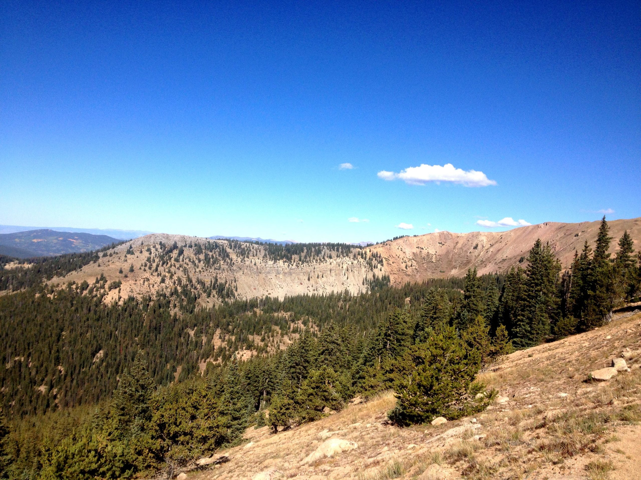 A panoramic view of a mountainous landscape featuring rugged hills and a clear blue sky. Pine trees dot the foreground, and the terrain transitions from greenery to rocky slopes in the background, showcasing the natural beauty of the area. CDT: Monarch Pass to Boss Lake / Hunt Lake Trail mountain bike trail.