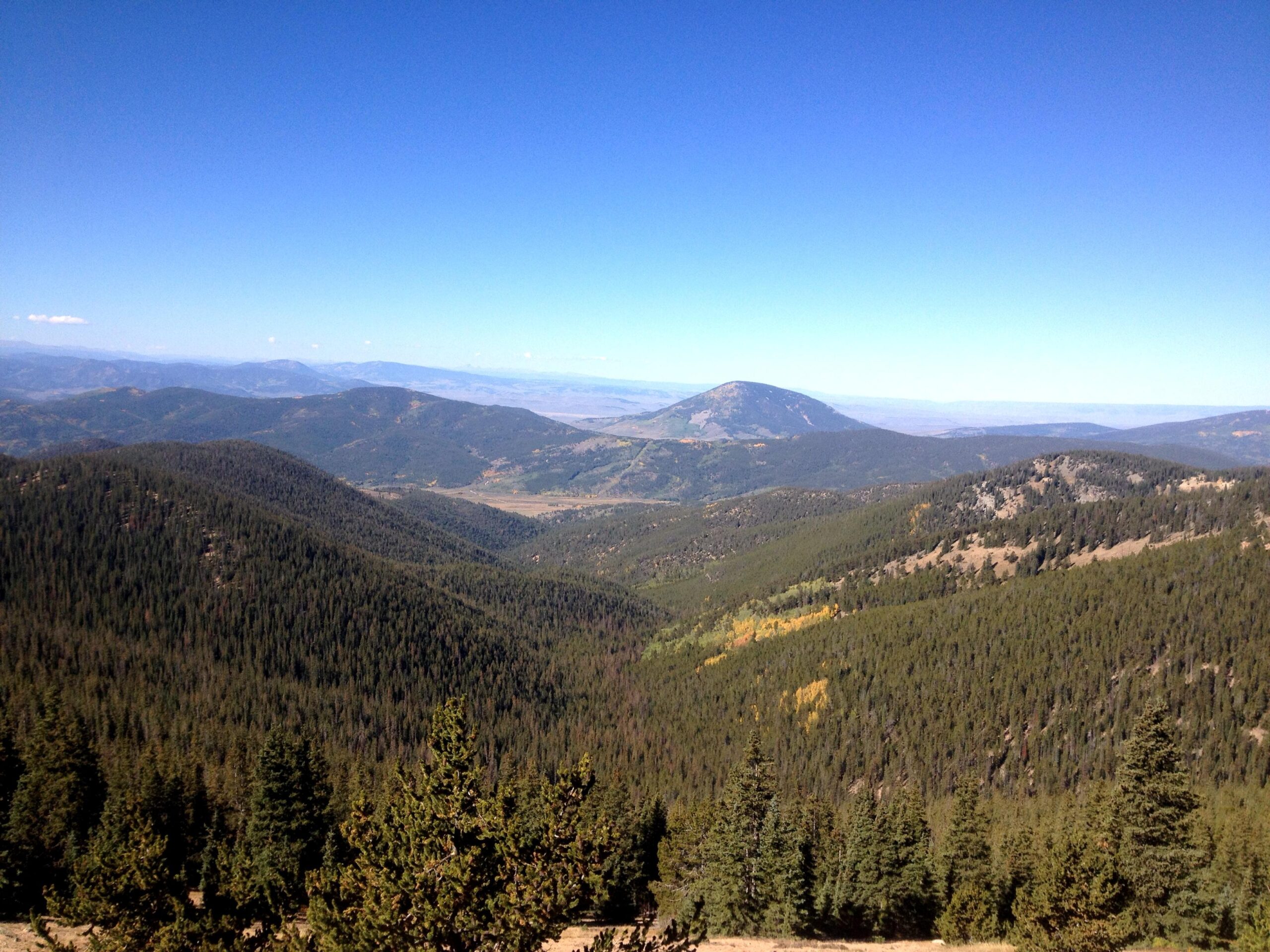 A panoramic view of rolling hills covered in lush green forests under a clear blue sky. In the distance, a prominent mountain peak rises, surrounded by valleys and more distant mountains. The landscape showcases a mix of greenery and hints of autumn colors. CDT: Monarch Pass to Boss Lake / Hunt Lake Trail mountain bike trail.