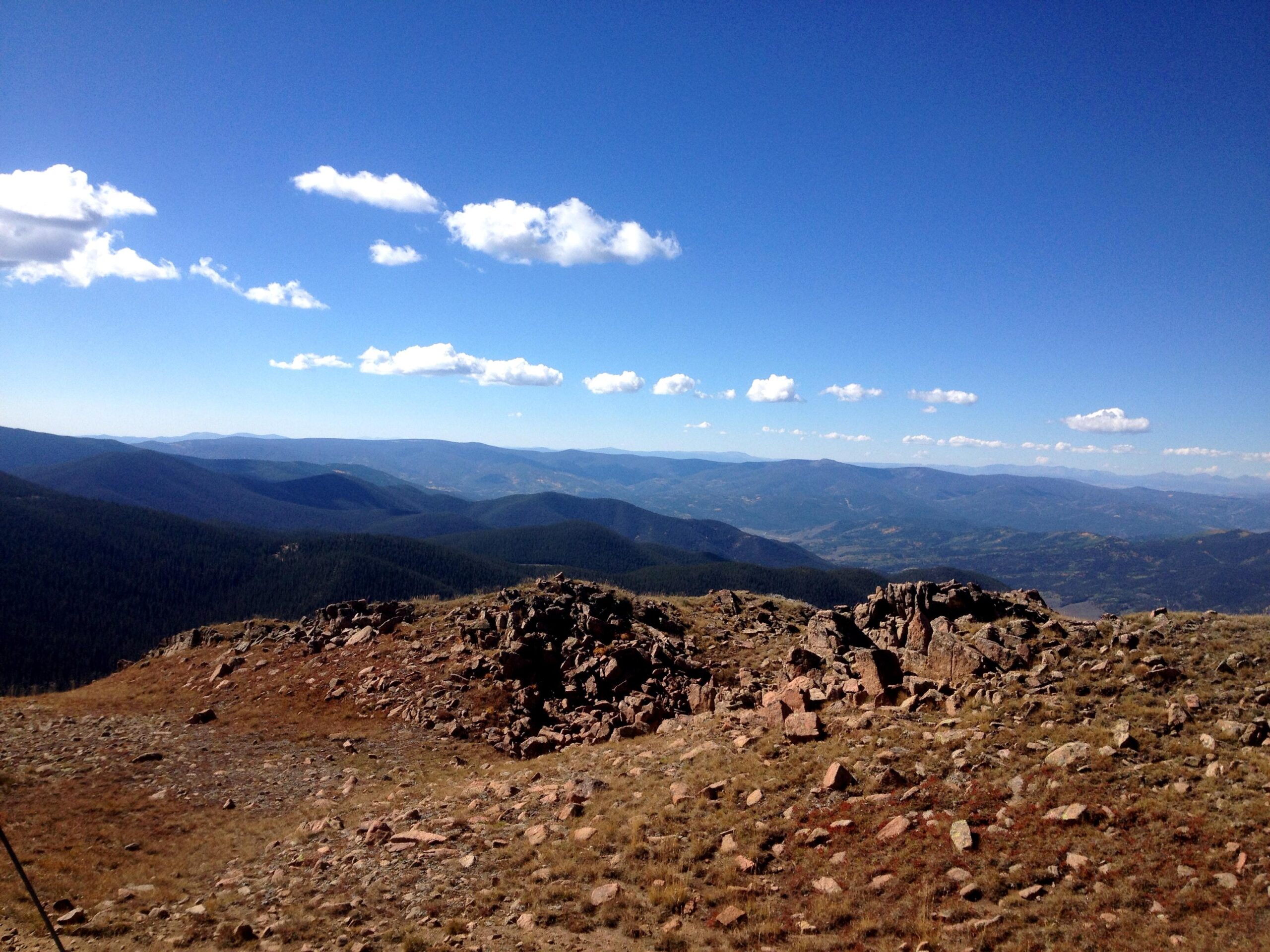 A scenic view of rolling mountains under a clear blue sky, featuring patches of clouds and rocky terrain in the foreground. The landscape showcases layers of green forests and distant peaks, creating a picturesque natural setting. CDT: Monarch Pass to Boss Lake / Hunt Lake Trail mountain bike trail.