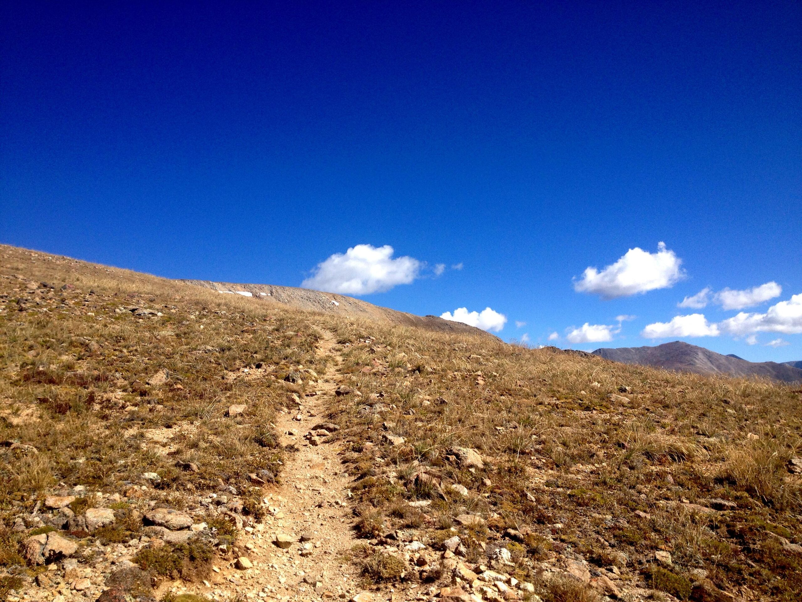 A winding dirt path leads upward through dry, grassy terrain under a bright blue sky with a few fluffy clouds, framed by distant mountains. CDT: Monarch Pass to Boss Lake / Hunt Lake Trail mountain bike trail.