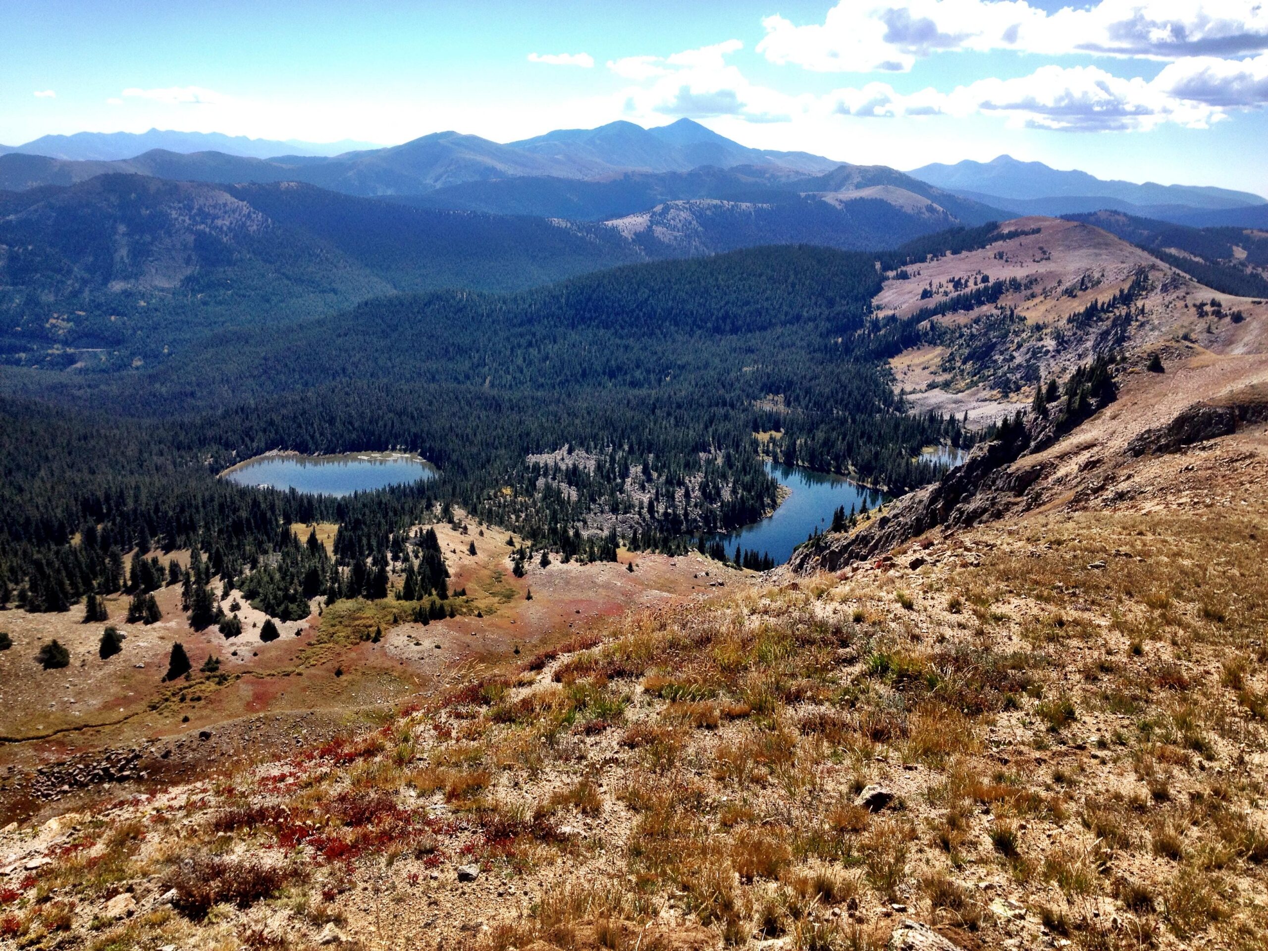 Aerial view of a mountainous landscape featuring two small lakes surrounded by dense forests and rocky terrain, with rolling hills and peaks in the background under a clear blue sky. CDT: Monarch Pass to Boss Lake / Hunt Lake Trail mountain bike trail.