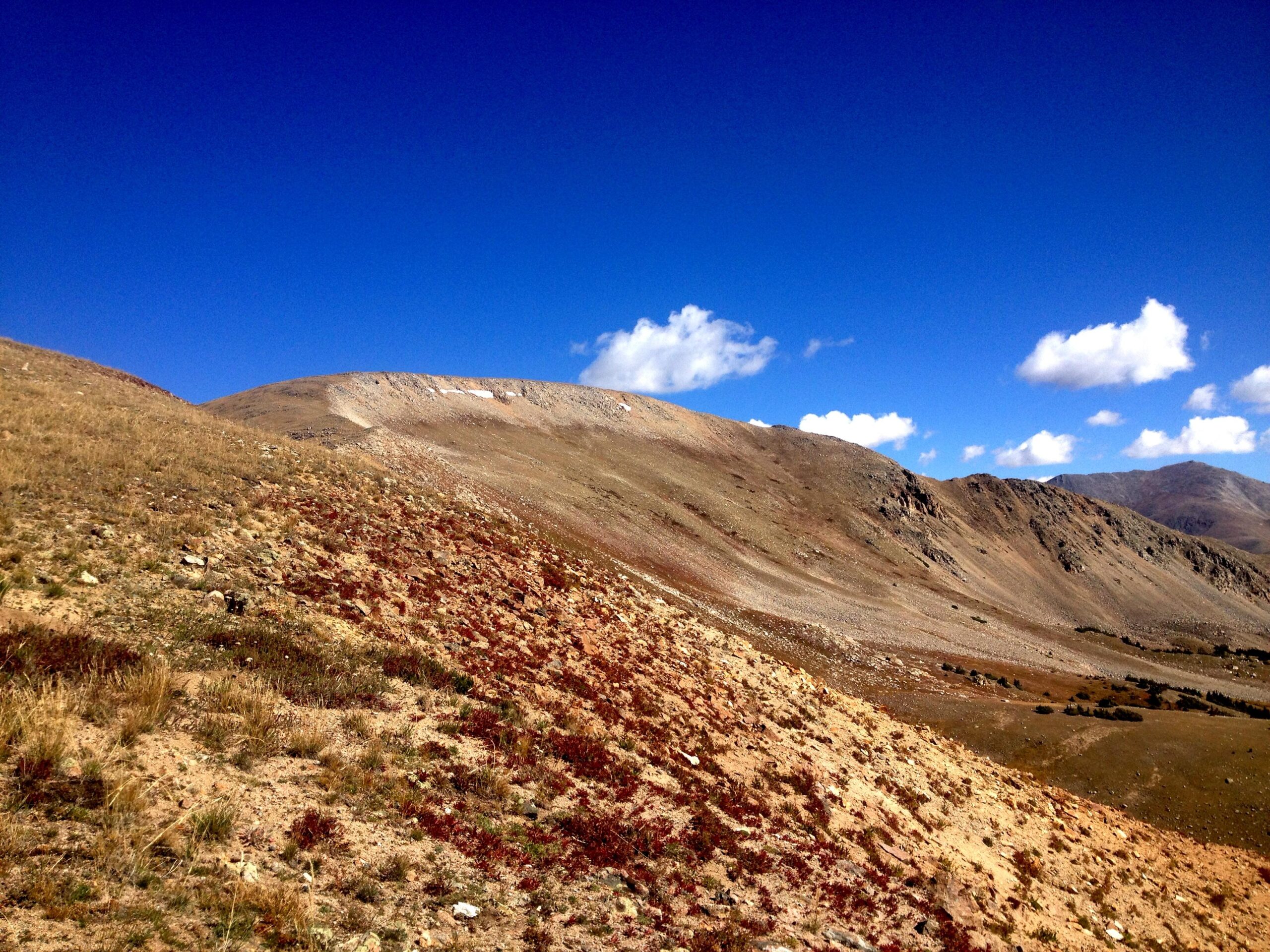 A scenic view of a mountainous landscape featuring rolling hills with dry grass and patches of red vegetation, under a clear blue sky with a few scattered clouds. CDT: Monarch Pass to Boss Lake / Hunt Lake Trail mountain bike trail.