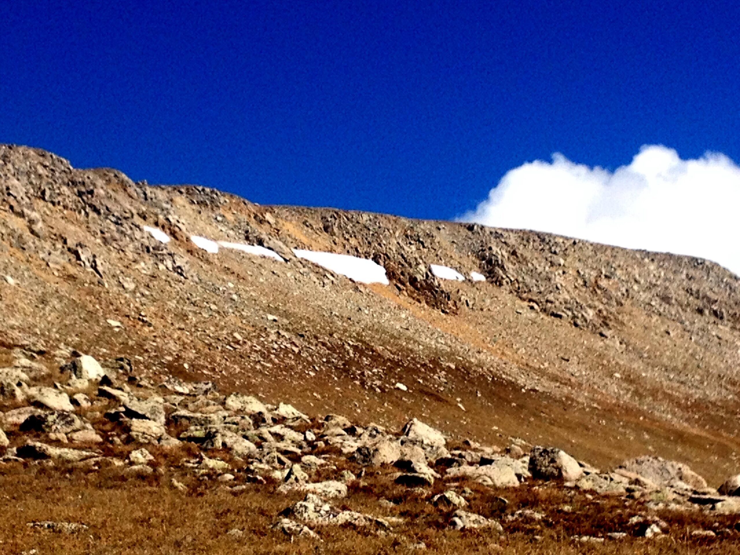 A rugged mountain landscape featuring a rocky slope with patches of snow and a deep blue sky dotted with clouds. The foreground consists of scattered boulders and dry grass, creating a natural, serene wilderness scene. CDT: Monarch Pass to Boss Lake / Hunt Lake Trail mountain bike trail.