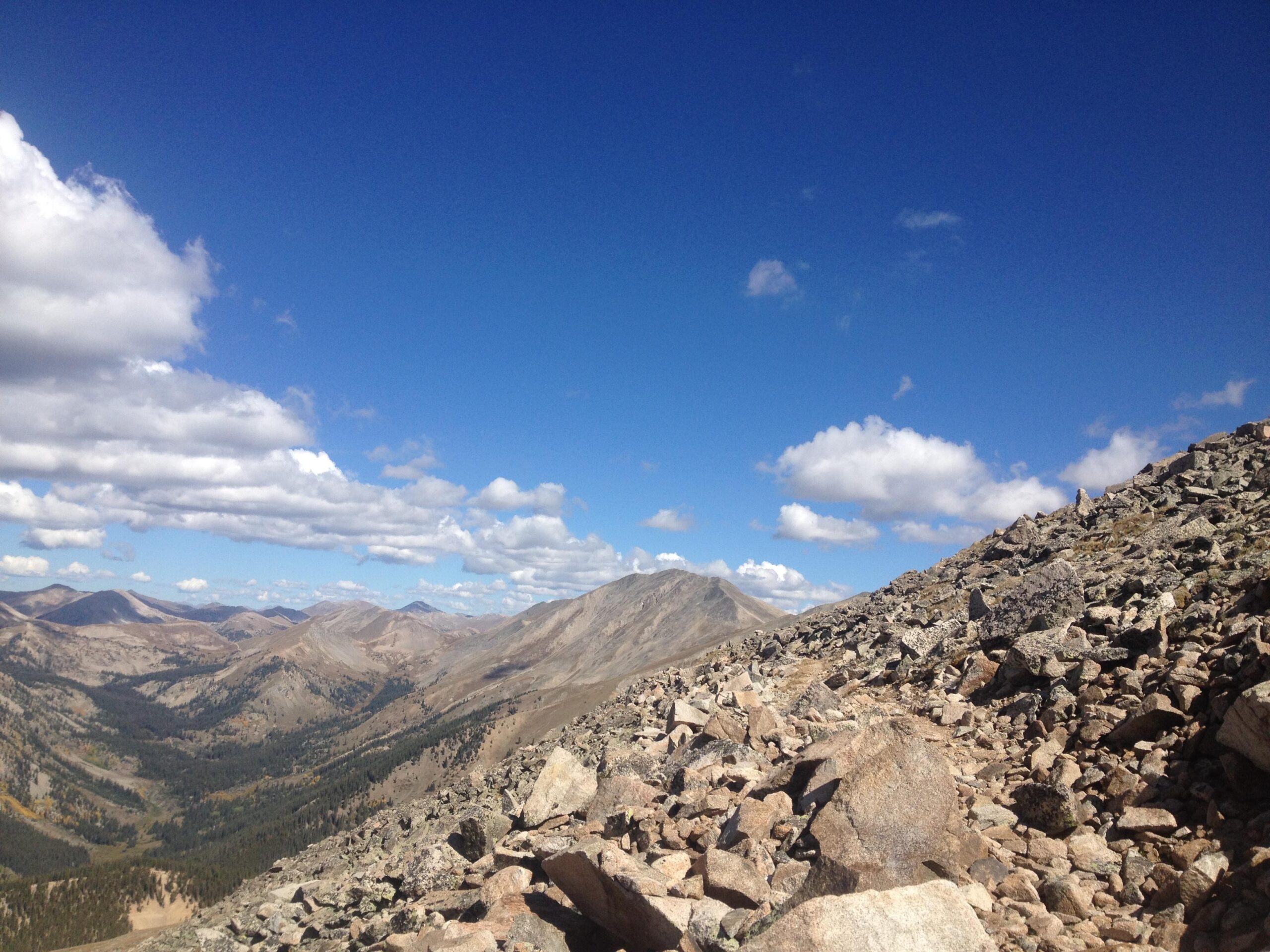 A panoramic view of a rocky mountain landscape under a clear blue sky, featuring scattered clouds. The foreground shows a rocky terrain leading to sloping mountains in the distance, with varied shades of brown and green indicating vegetation and geological diversity. The scene captures a sense of natural beauty and vastness. CDT: Monarch Pass to Boss Lake / Hunt Lake Trail mountain bike trail.