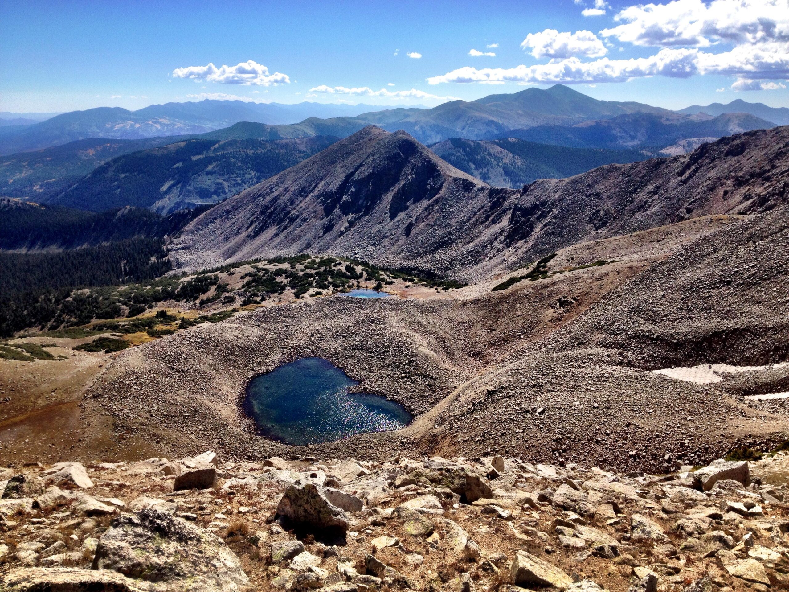 A panoramic view of rugged mountain terrain featuring a small, clear pond nestled among rocky outcrops. The landscape is dotted with patches of green vegetation, with peaks in the background under a bright blue sky scattered with fluffy white clouds. Sunlight reflects off the water's surface, highlighting the natural beauty of the area. CDT: Monarch Pass to Boss Lake / Hunt Lake Trail mountain bike trail.