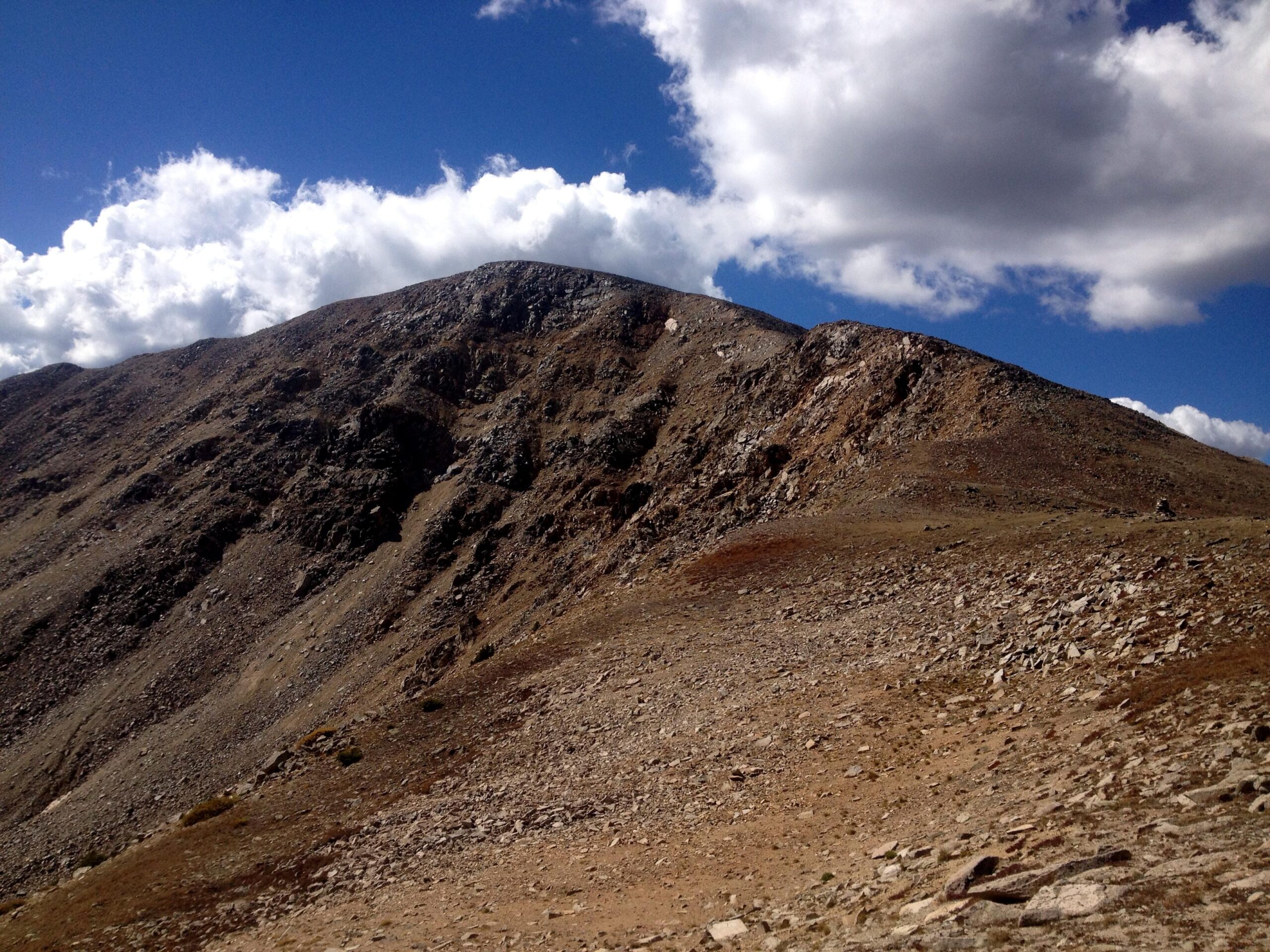 A rugged mountain landscape featuring a rocky slope leading up to a peak, under a bright blue sky with scattered clouds. The foreground shows a barren terrain with sparse vegetation and scattered stones. CDT: Monarch Pass to Boss Lake / Hunt Lake Trail mountain bike trail.