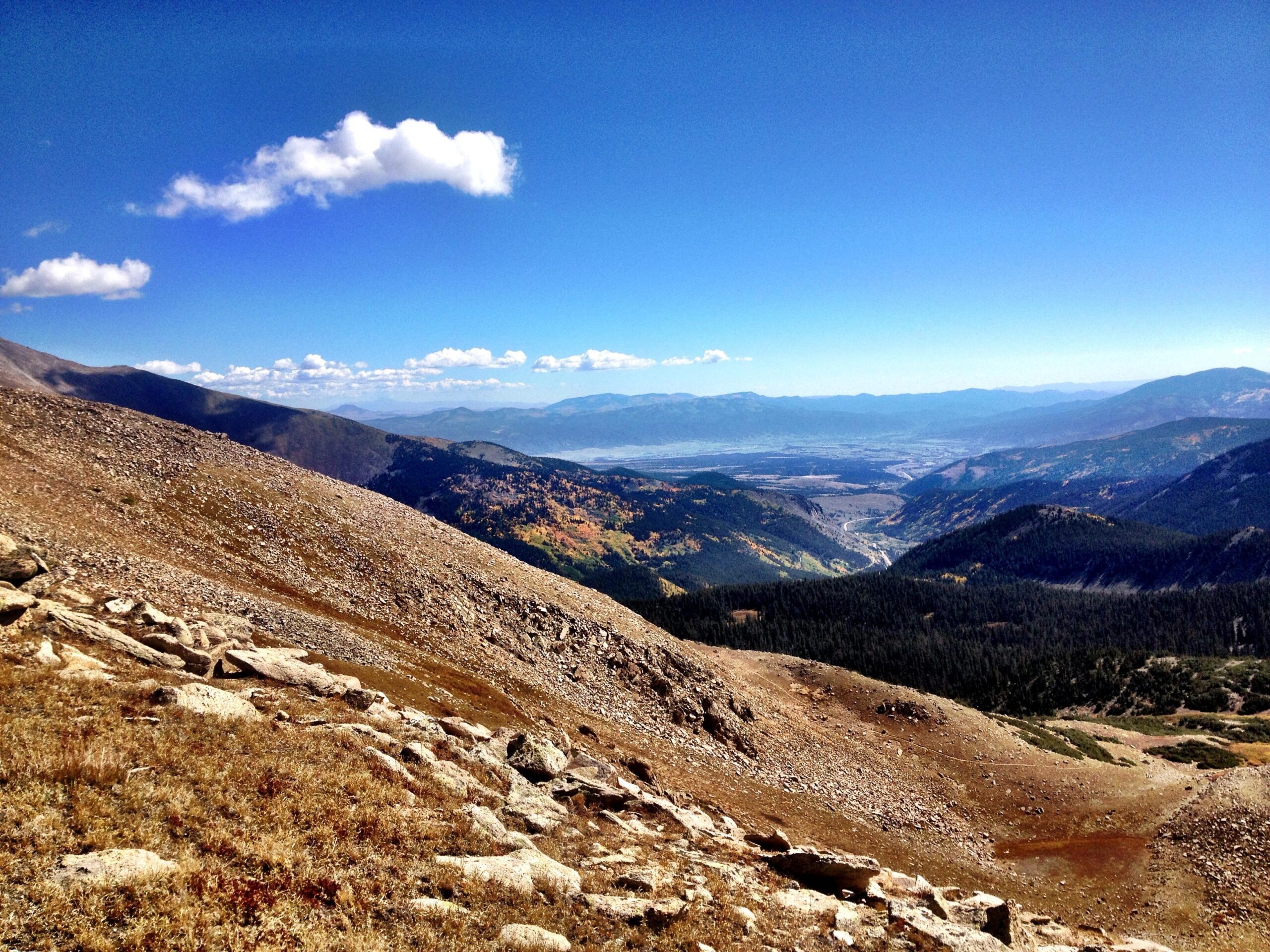A panoramic view of a mountainous landscape featuring rolling hills and valleys, under a bright blue sky dotted with a few white clouds. The foreground shows rocky terrain with sparse vegetation, while the background reveals distant mountains and valleys with patches of greenery, suggesting a mix of forests and fields. The sunlight enhances the natural colors, creating a serene and picturesque outdoor scene. CDT: Monarch Pass to Boss Lake / Hunt Lake Trail mountain bike trail.