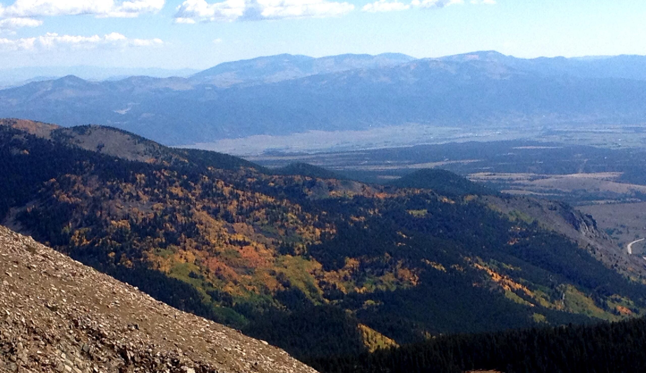 A panoramic view of a mountainous landscape, showcasing rolling hills covered in trees, some displaying autumn colors of gold and green. In the distance, a range of mountains stretches across the horizon under a partly cloudy sky. The foreground features rocky terrain leading into the lush hills below. CDT: Monarch Pass to Boss Lake / Hunt Lake Trail mountain bike trail.