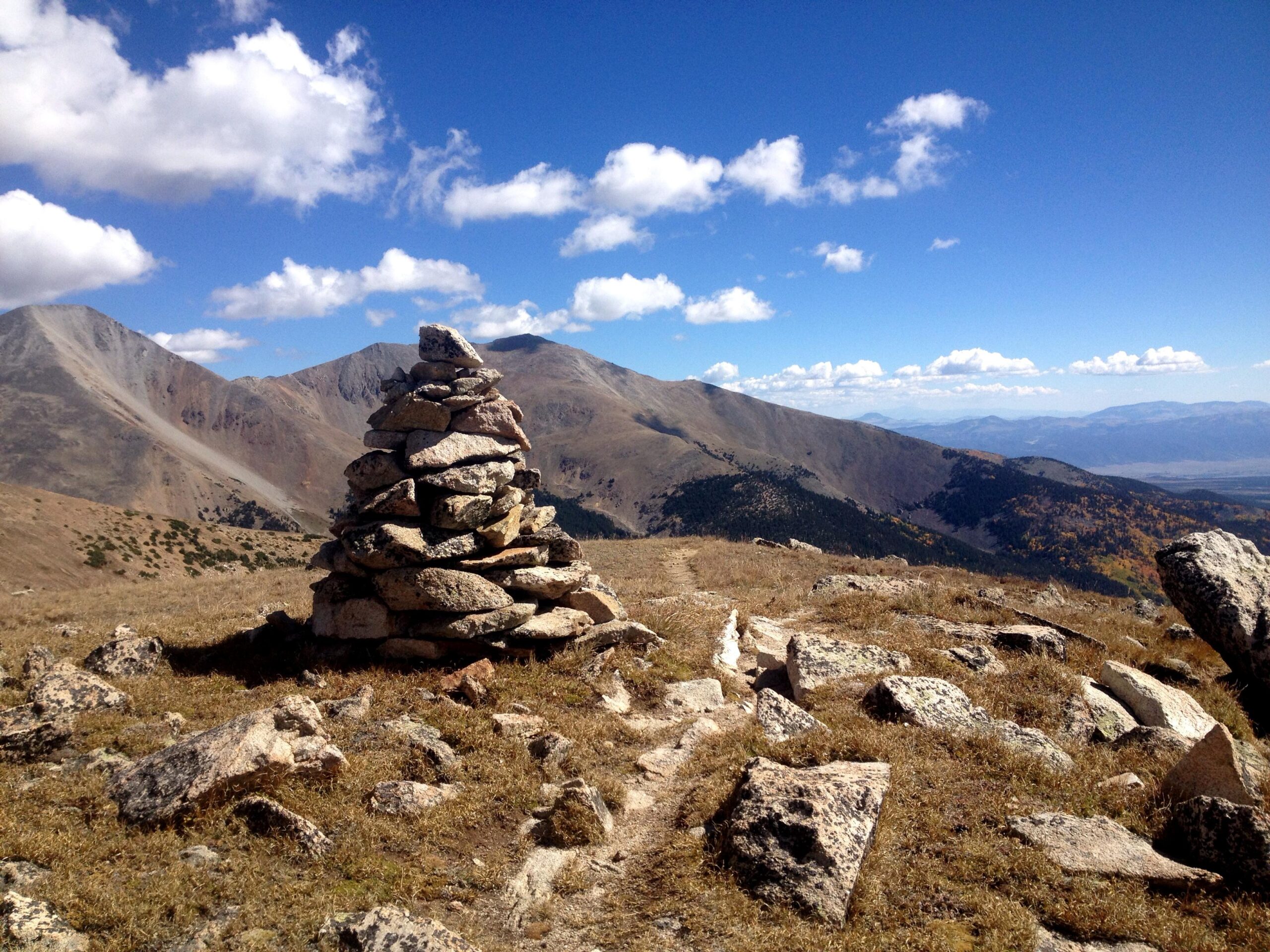 A stone cairn stands prominently on a grassy mountainside, surrounded by rocky terrain and distant peaks under a bright blue sky filled with fluffy white clouds. The landscape features a mix of rugged mountains and open meadows, showcasing the natural beauty of the area. CDT: Monarch Pass to Boss Lake / Hunt Lake Trail mountain bike trail.