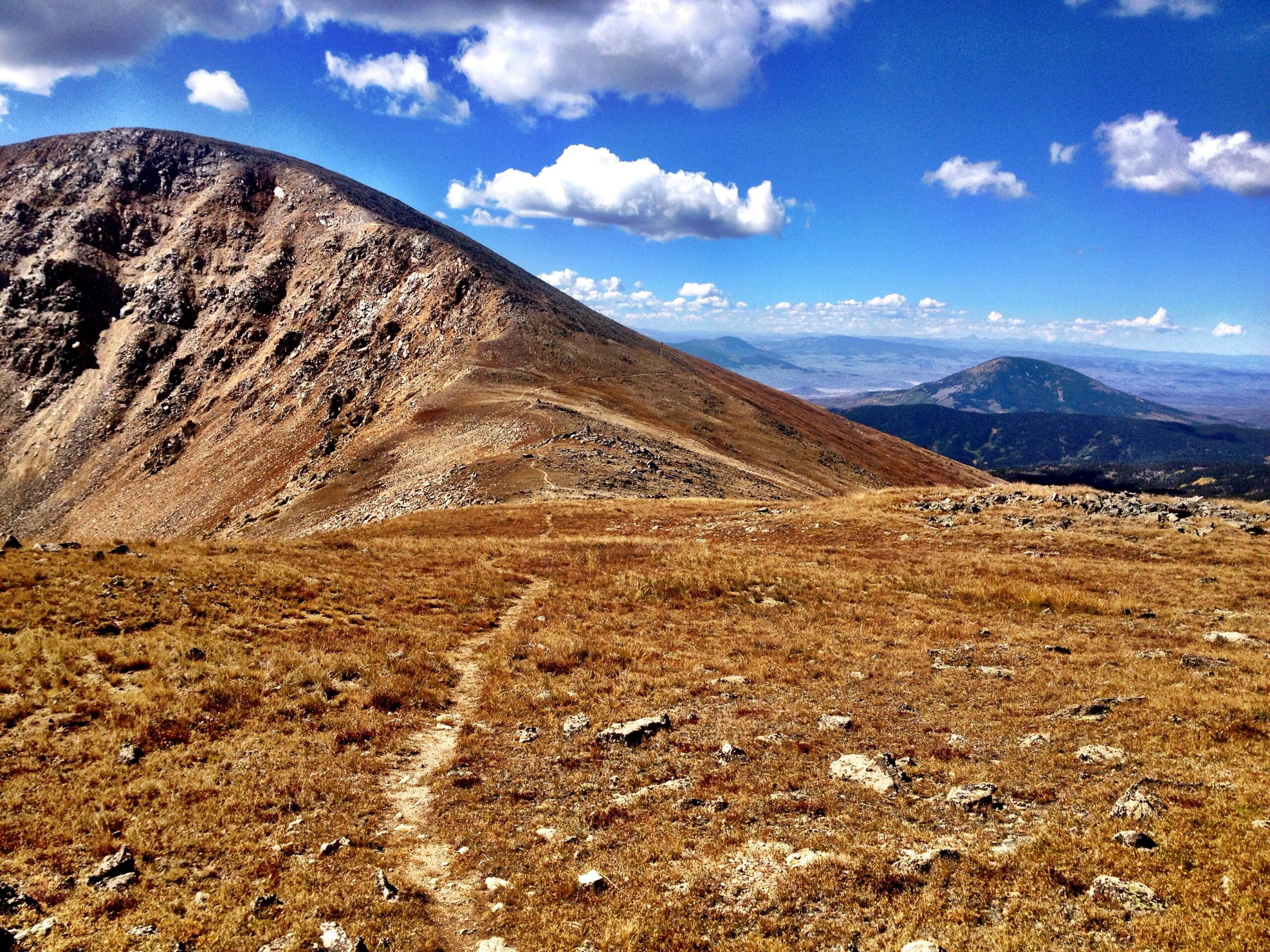 A panoramic view of a mountainous landscape featuring rolling hills with rocky terrain and patches of golden grass under a bright blue sky adorned with fluffy clouds. A narrow dirt path winds through the foreground, leading towards the peaks in the background. CDT: Monarch Pass to Boss Lake / Hunt Lake Trail mountain bike trail.