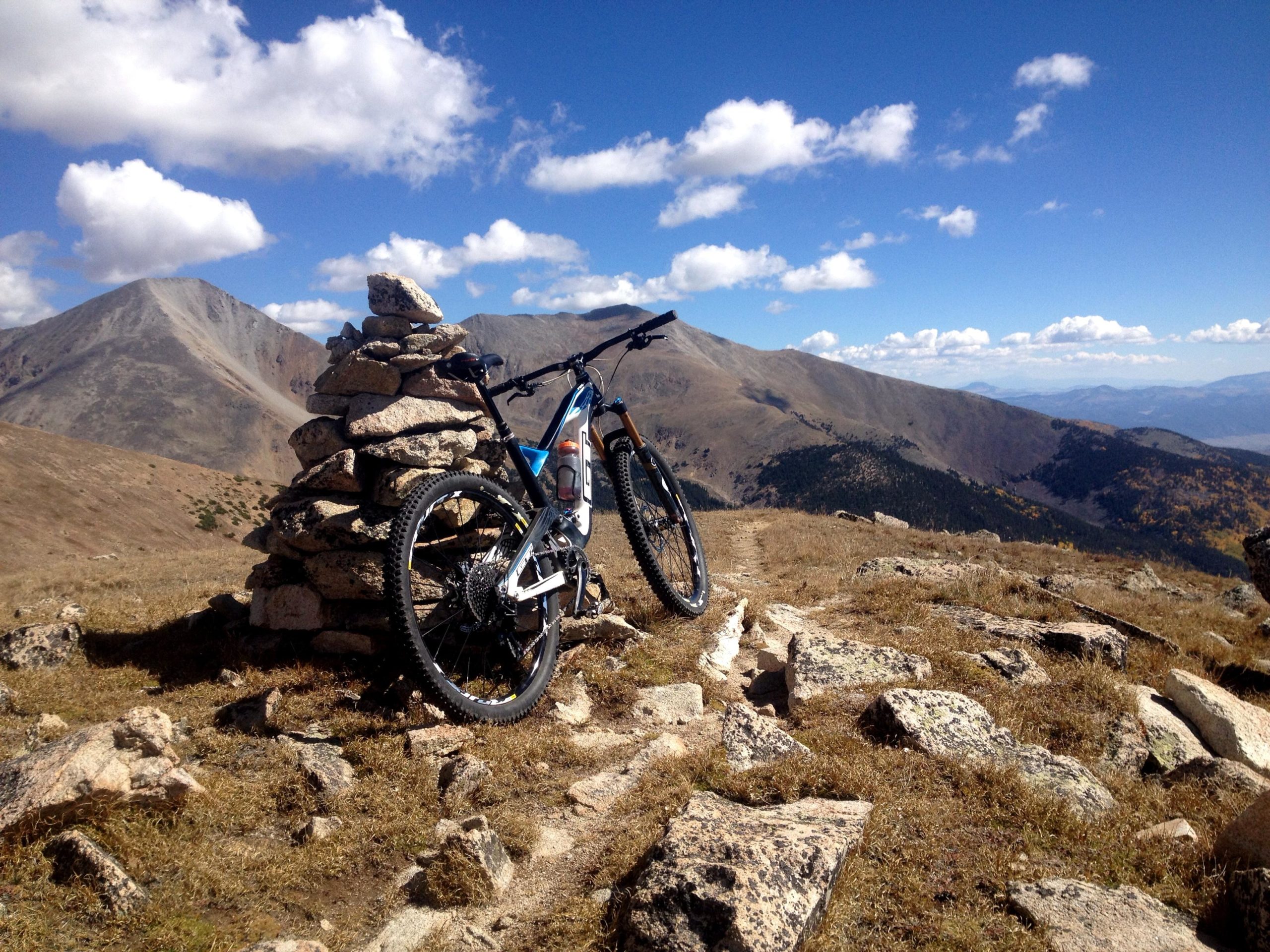 A mountain bike resting against a stone cairn on a mountainous terrain, with expansive views of rolling hills and a blue sky dotted with clouds. The scene captures the beauty of outdoor adventure in a rugged landscape. CDT: Monarch Pass to Boss Lake / Hunt Lake Trail mountain bike trail.