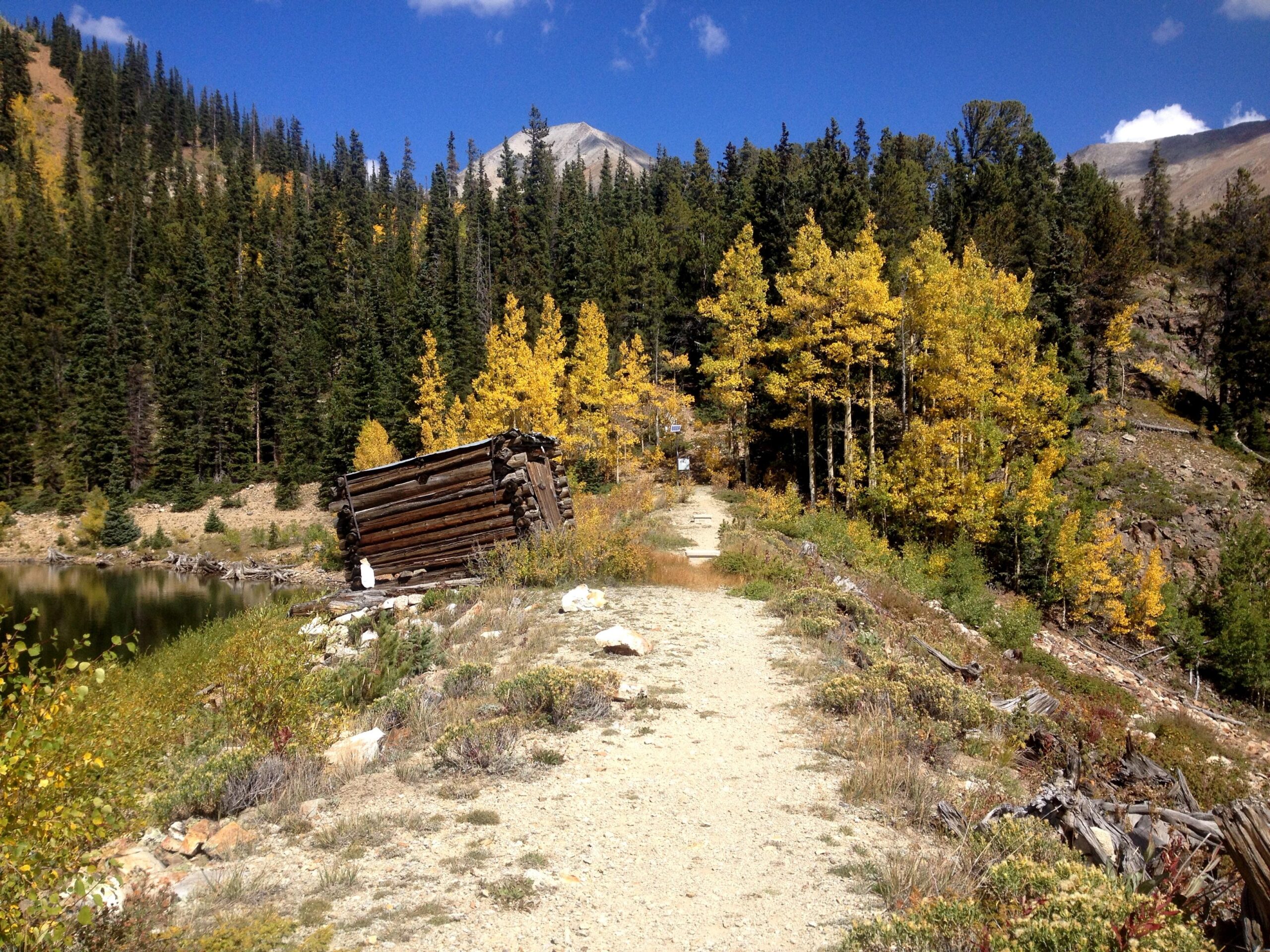 A winding path leading through a mountainous landscape, bordered by vibrant autumn trees with yellow leaves. An old, leaning wooden cabin sits near the edge of a serene body of water, surrounded by lush greenery and rocky terrain. The sky is clear with a few clouds, and distant mountains rise in the background. CDT: Monarch Pass to Boss Lake / Hunt Lake Trail mountain bike trail.