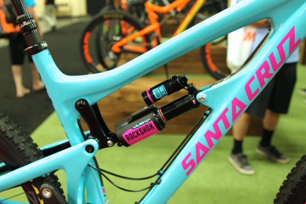 Santa Cruz Nomad Carbon 27.5: Close-up view of the shock system on a blue Santa Cruz mountain bike frame, featuring a RockShox shock absorber. The frame displays bold pink lettering of the "SANTA CRUZ" logo, with a blurred background of other bicycles and a green floor.