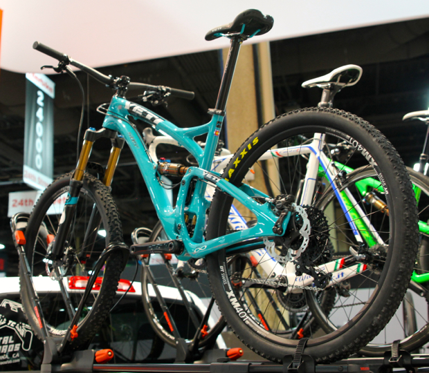 A turquoise mountain bike displayed on a rack at an exhibition, featuring thick tires and a sleek frame design, with other bicycles in the background.