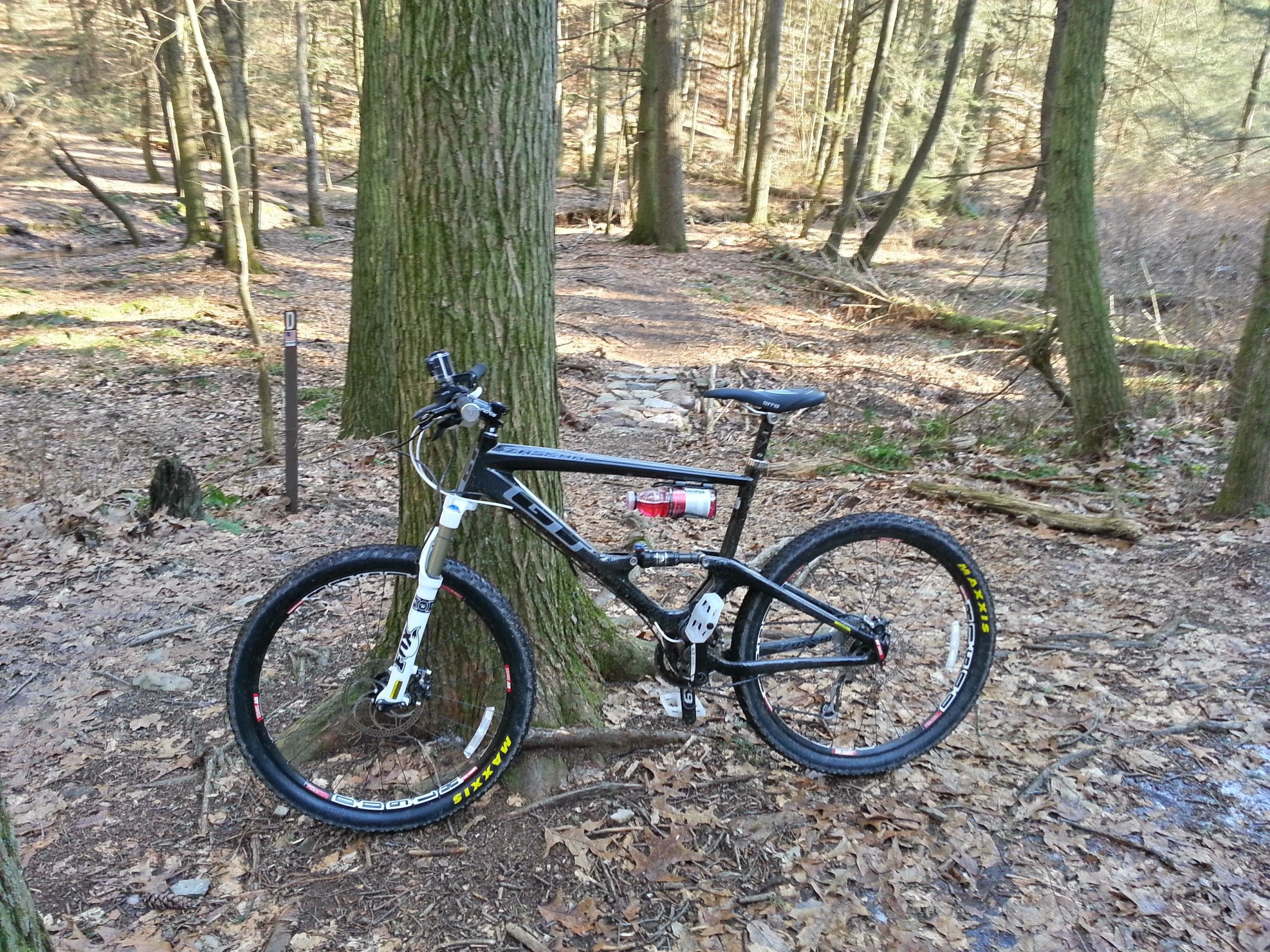 A black and white mountain bike leaning against a tree in a forested area. The ground is covered with fallen leaves, and a trail can be seen in the background. A signpost is partially visible, indicating a designated trail marker. The scene is peaceful and captures the essence of outdoor cycling. Swatara State Park mountain bike trail.