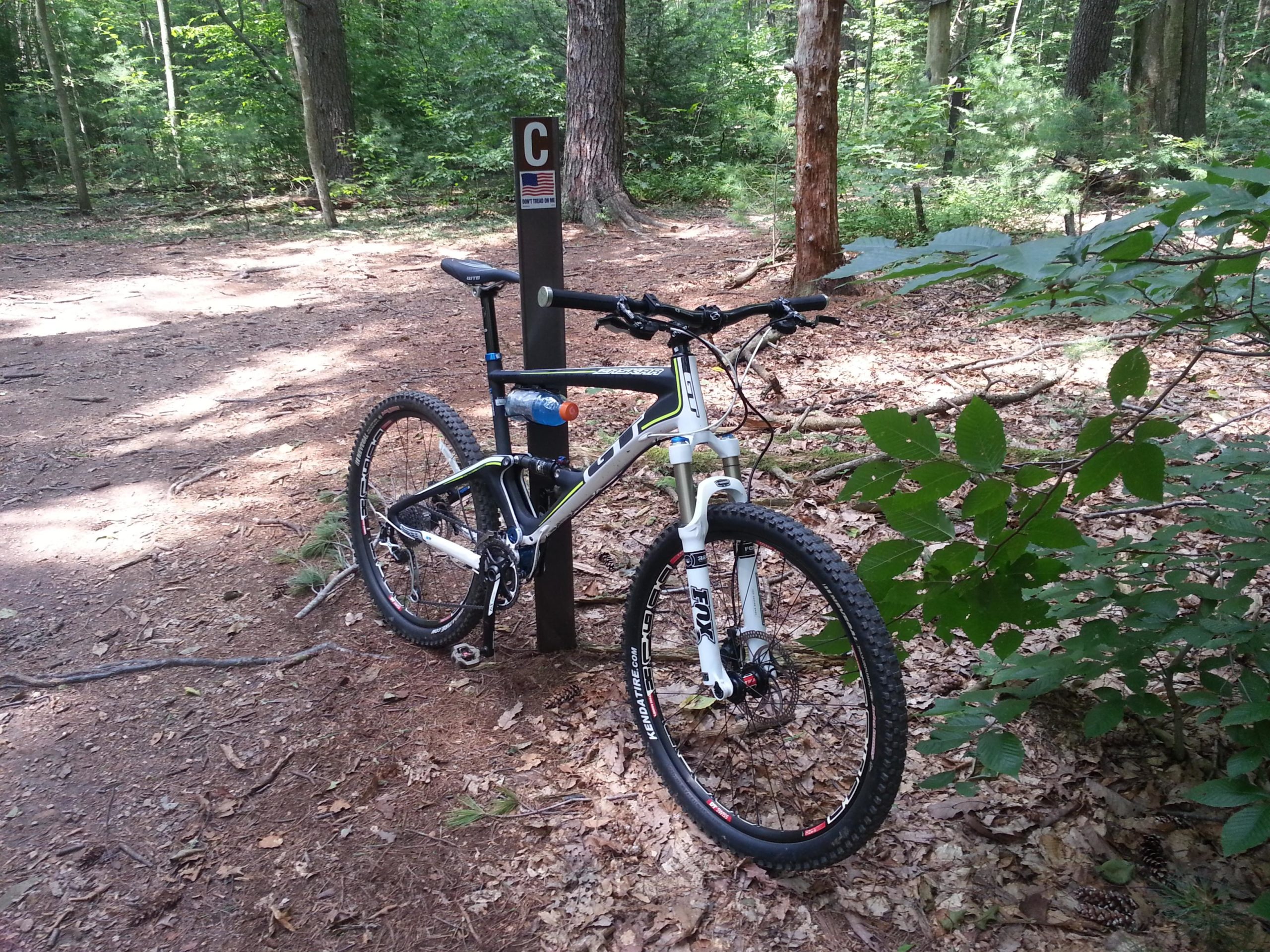 Mountain bike leaning against a trail marker in a wooded area, with a well-trodden dirt path visible. Sunlight filters through the trees, highlighting the greenery and fallen leaves on the ground. The trail marker is labeled "C" and features a small American flag sticker. Swatara State Park mountain bike trail.
