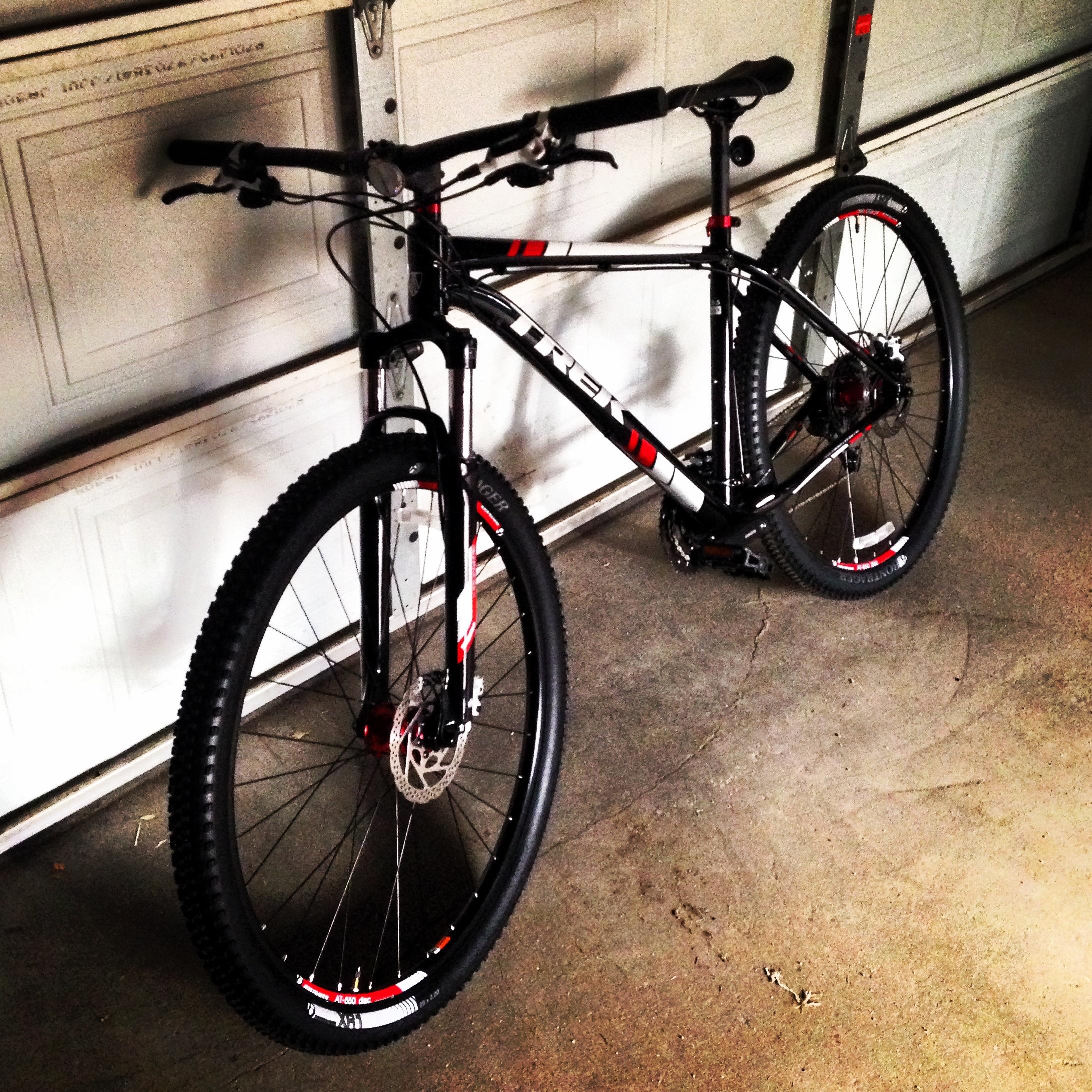 Trek X-Caliber 6: A black and red mountain bike leaning against a garage wall, with thick tires and disc brakes, positioned on a concrete floor.