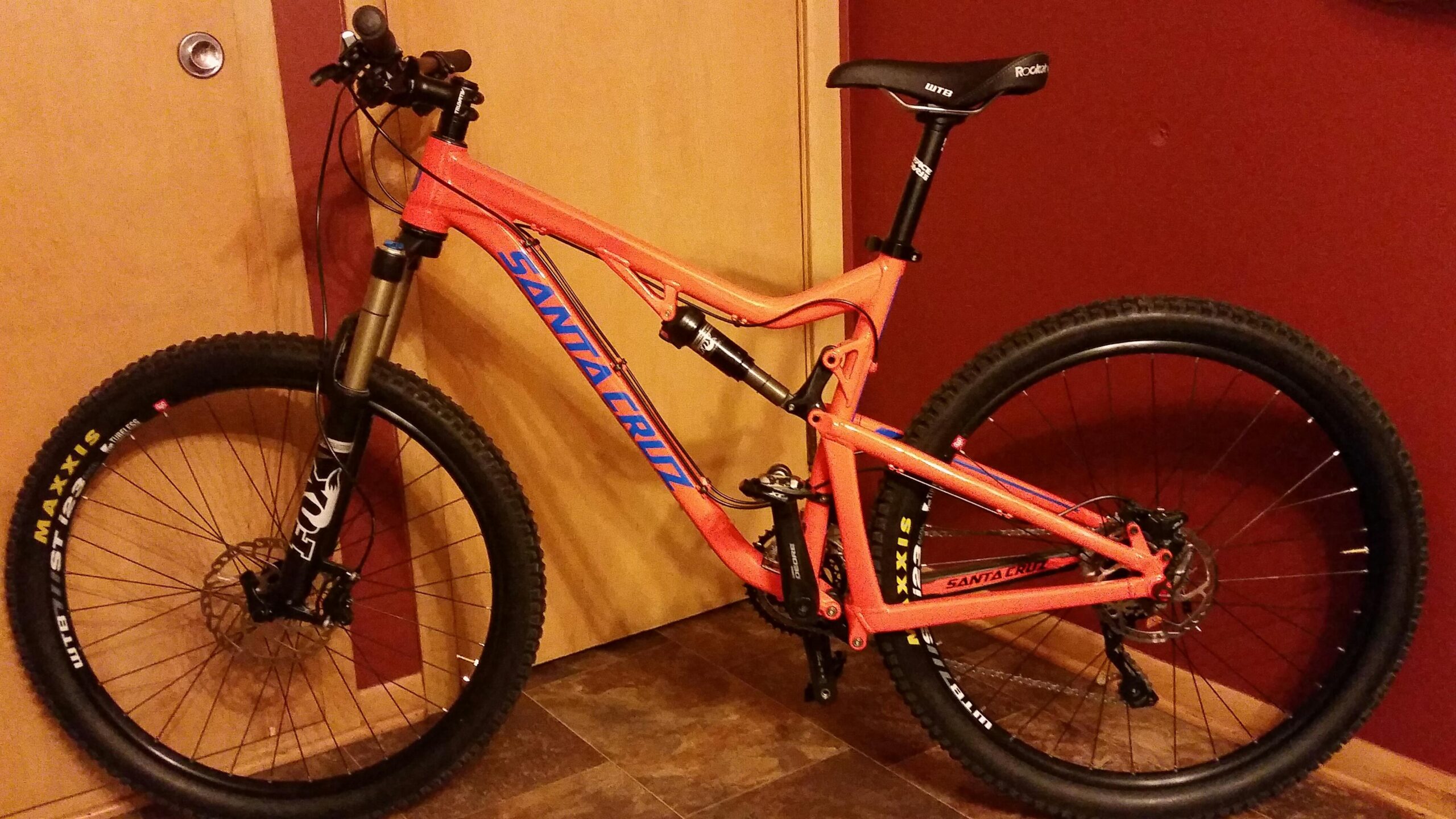 Santa Cruz 5010: A bright orange mountain bike with distinctive blue branding, featuring a full suspension system and thick tires designed for off-road terrain. The bike is positioned upright against a wooden door and wall, showcasing its sleek frame and mechanical components.