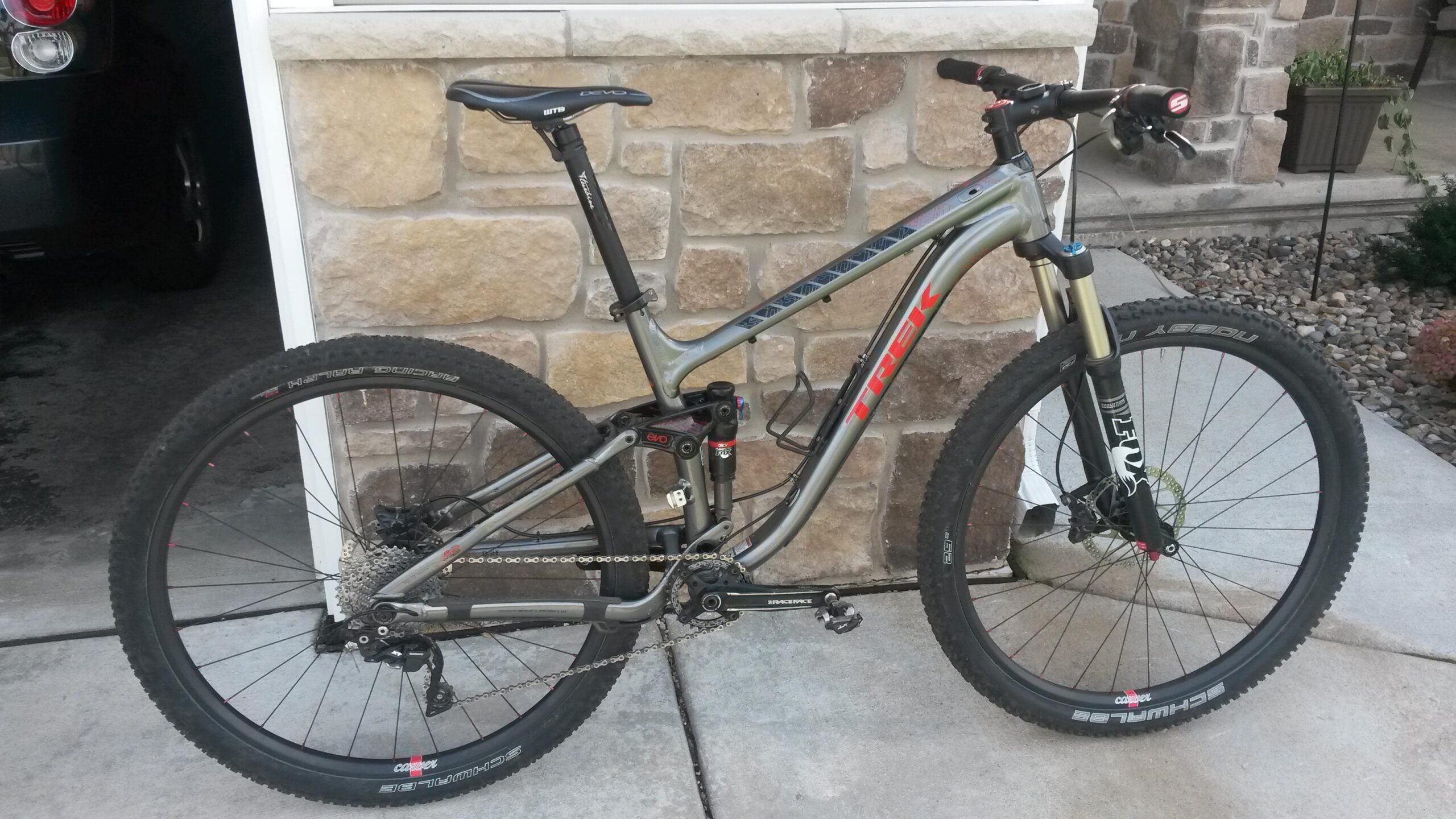 Trek Fuel EX 8 29: A silver and black mountain bike leaning against a stone wall, featuring thick tires, a sturdy frame, and various gear components visible. The bike includes a suspension system and is positioned on a concrete surface with a landscaped area in the background.