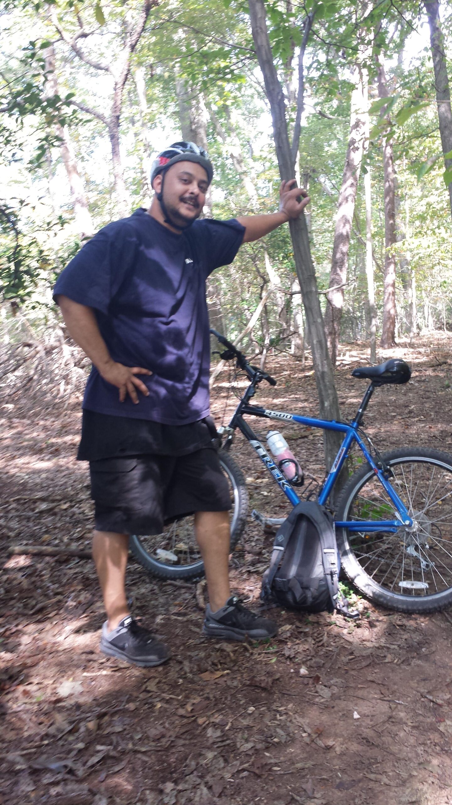 A person wearing a helmet stands beside a blue mountain bike in a wooded area, smiling and posing with one hand on a tree and the other on their hip. The ground is covered with leaves, and trees surround the scene, creating a natural outdoor setting. A backpack is leaning against the bike, and a water bottle is attached to the bike frame. Clopper Lake mountain bike trail.