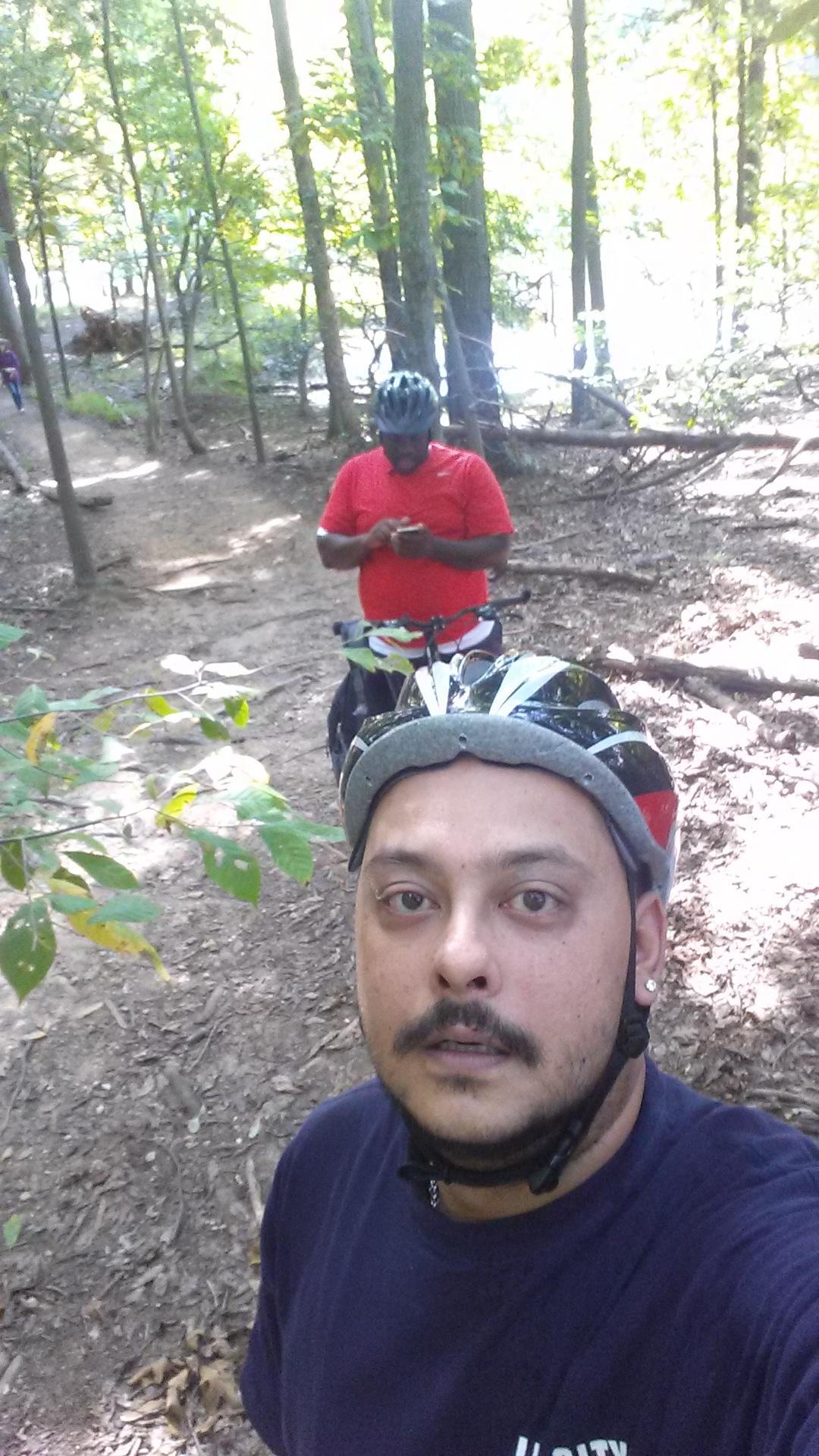 A person wearing a helmet takes a selfie while biking on a wooded trail. In the background, another individual in a red shirt appears to be checking their phone. The scene is set in a lush green forest with trees and a dirt path visible. Clopper Lake mountain bike trail.