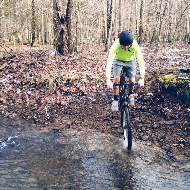 A person wearing a bright green long-sleeve shirt and a helmet is riding a mountain bike over a shallow stream in a forested area. The ground is muddy and surrounded by trees, indicating an outdoor adventure setting. The cyclist is mid-jump, with the front wheel lifted above the water. Snake Root mountain bike trail.