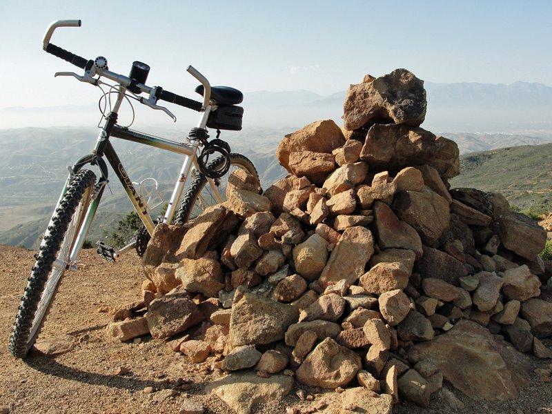 Giant Rincon: A mountain bike leaning against a pile of stacked rocks on a rocky hillside, with a scenic view of mountains and valleys in the background under a clear blue sky.