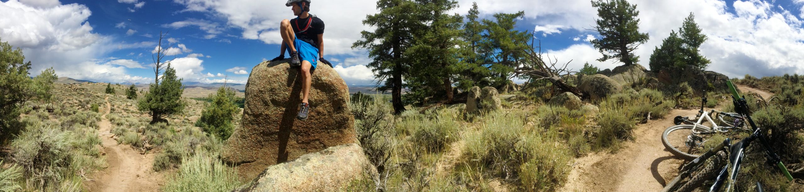 A mountain biker sits on a large boulder overlooking a scenic landscape with sparse vegetation and scattered rocks. In the foreground, a white bicycle lies on its side along a dirt trail winding through the area. The sky is partly cloudy with hints of blue and distant mountains visible in the background. Hartman Rocks mountain bike trail.