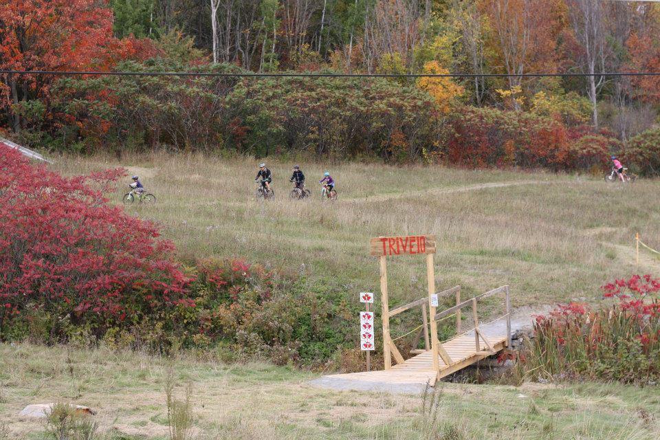A group of five mountain bikers riding on a grassy trail surrounded by colorful autumn foliage. In the foreground, there is a wooden bridge with a sign that reads "TRIVET." The scene captures the vibrant greens, reds, and yellows of fall. Sentier #1 mountain bike trail.