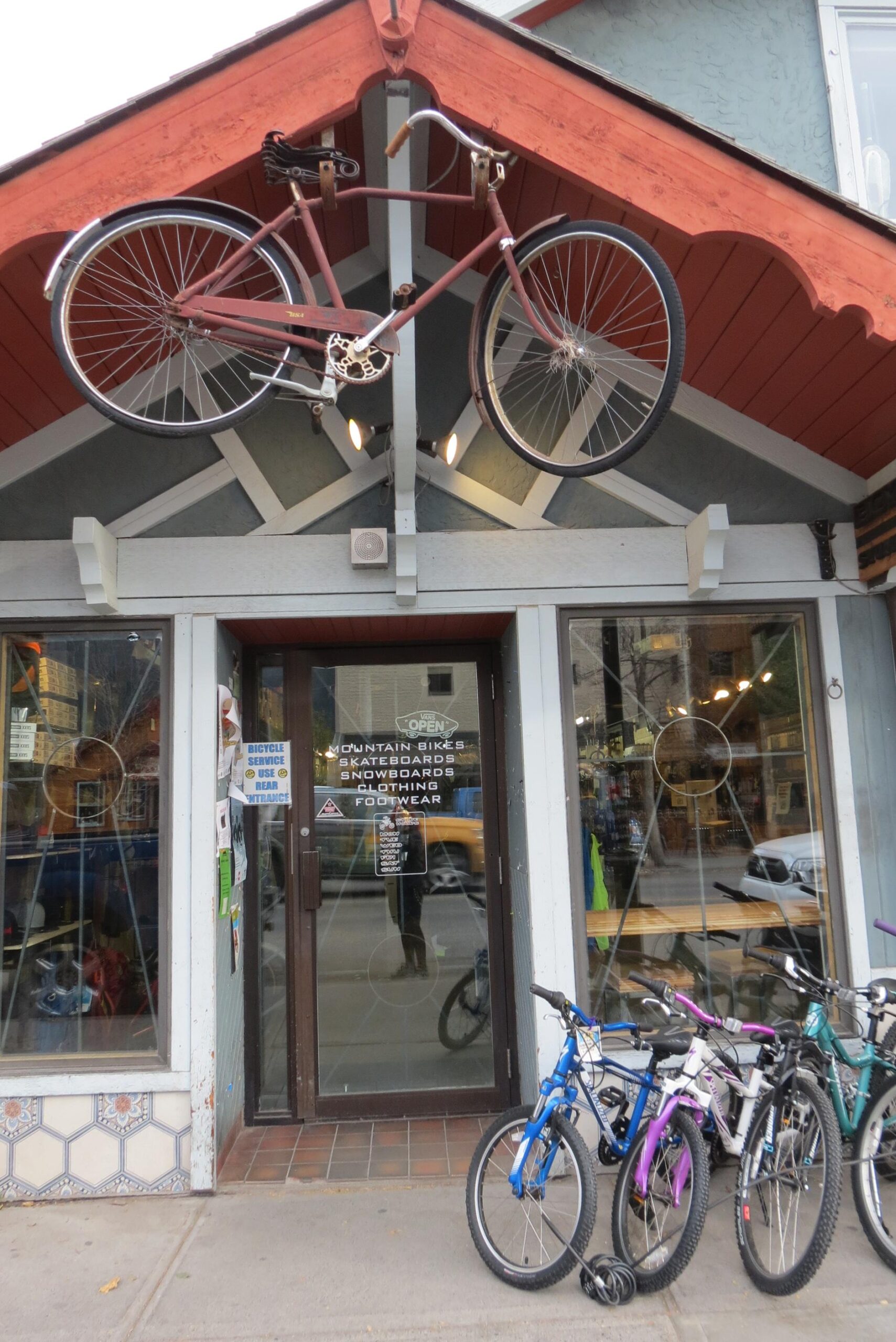 An image of a storefront featuring a bicycle mounted on the roof above the entrance. The door shows several signs indicating the shop is open and sells mountain bikes, skateboards, snowboards, clothing, and footwear. There are additional bicycles parked outside the store. The architecture includes decorative wooden elements and tiled flooring.