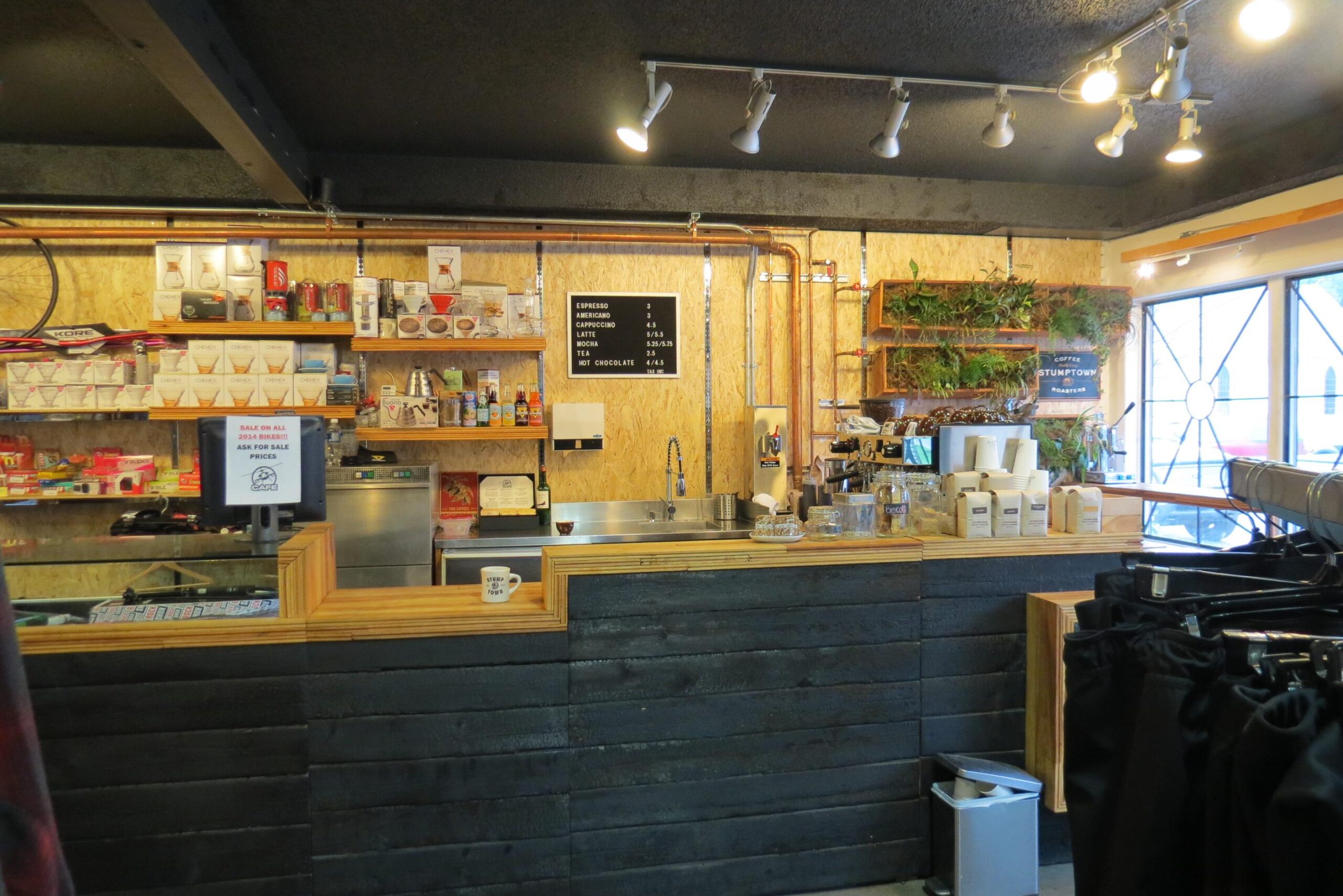 A cozy café counter featuring a variety of drinks and snacks for sale. The wooden bar is complemented by a rustic backdrop, with shelves displaying coffee supplies and beverages. A chalkboard menu lists prices for espresso, cappuccino, and hot chocolate. The area is well-lit with track lighting, contributing to a warm atmosphere. A few potted plants add a touch of greenery, while a clothing rack with black garments is visible in the foreground.