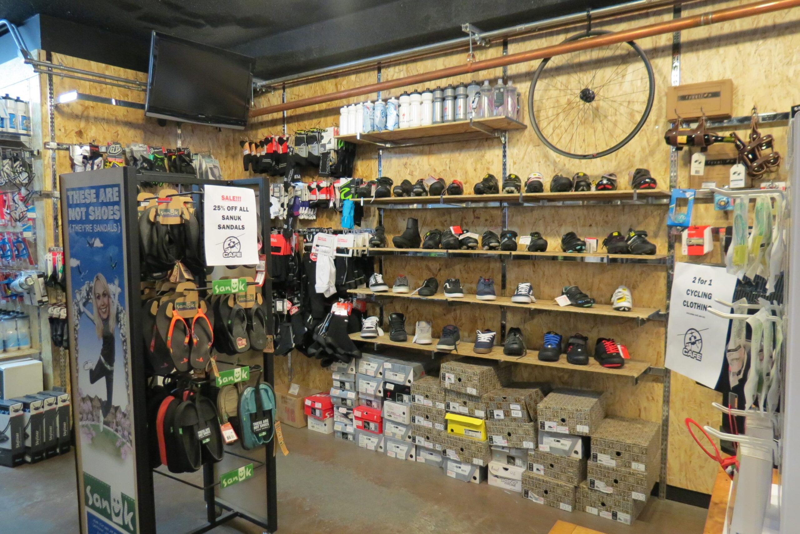 Store interior featuring racks of various cycling shoes and sandals. A display advertises a sale on Sanuk sandals, with colorful cycling gear hanging on the walls. Shelves are stocked with a variety of shoe boxes, and a television is mounted in the background.
