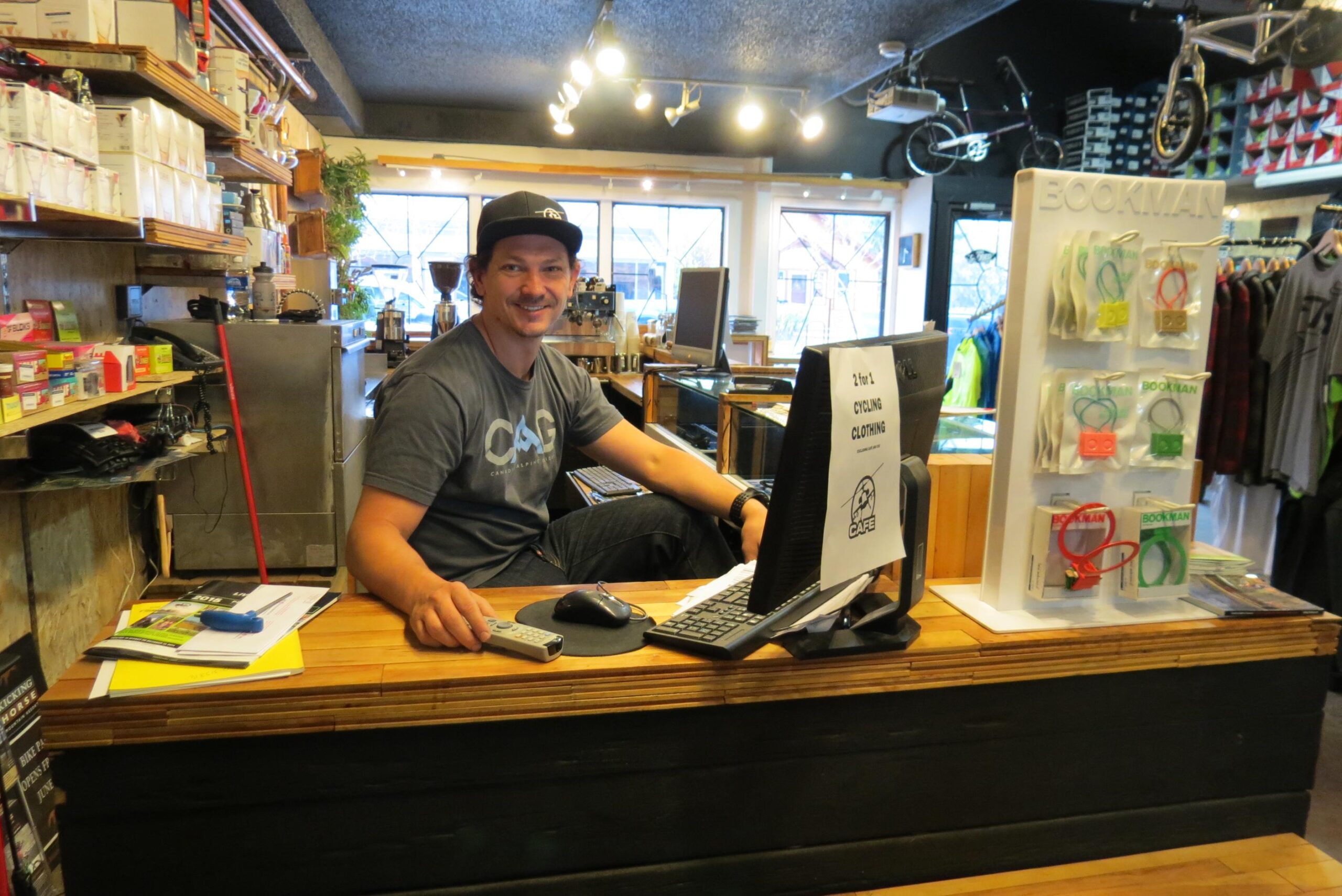 A man sitting at a wooden counter in a retail store, smiling while looking at the camera. He is wearing a gray t-shirt and a black cap. Behind him, shelves are stocked with various products, including cycling clothing and accessories. A computer monitor and keyboard are positioned on the counter, with a sign that reads "2 for 1 CYCLING CLOTHING." The shop interior features warm lighting and displays of merchandise.