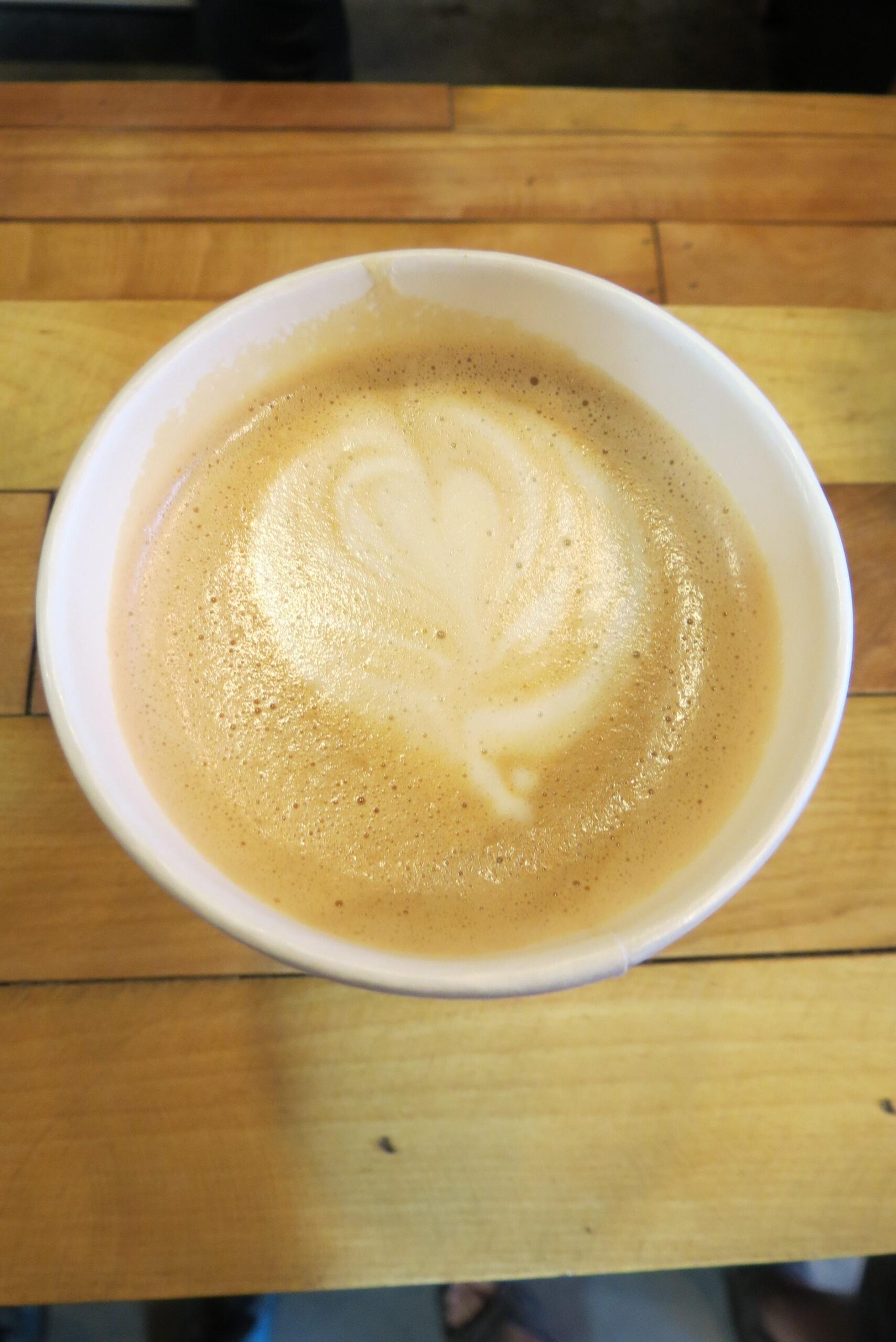 A close-up view of a latte in a white cup, featuring a heart-shaped latte art design on the frothy surface. The cup is placed on a wooden table, showcasing its warm tones.
