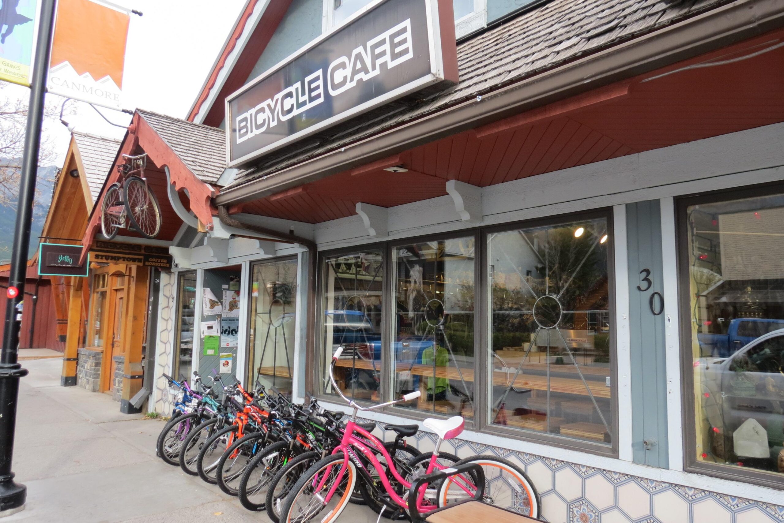 A street view of a bicycle cafe with a large sign reading "BICYCLE CAFE" above the entrance. Multiple bicycles are parked outside, showcasing a variety of colors, including pink, orange, and black. The cafe features large windows that reveal wooden furniture inside. Nearby, a sign indicating "Stubb's Coffee" is visible, along with a decorative wooden building on the left.