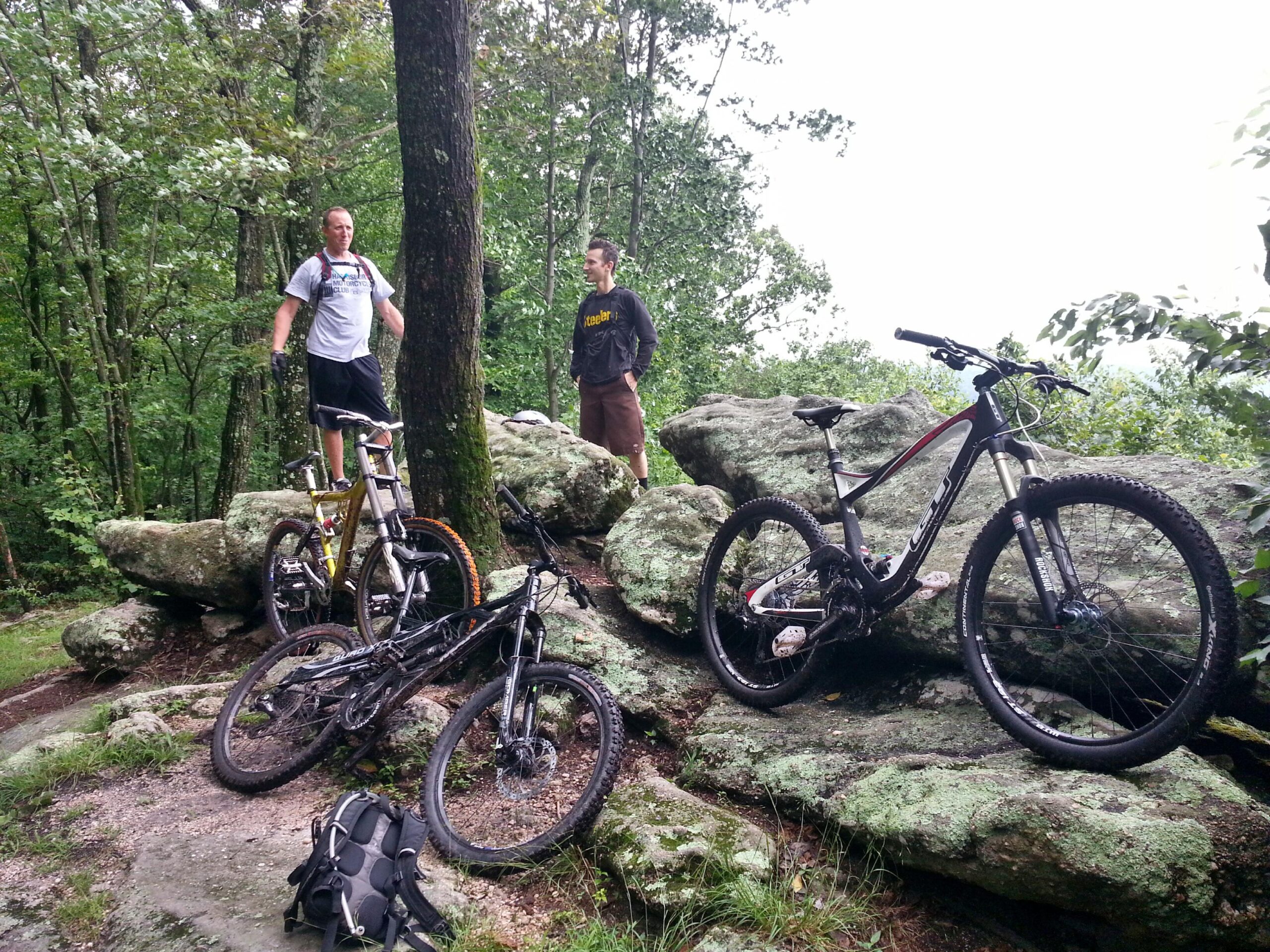 Two mountain bikers are standing near a rocky outcrop in a wooded area, surrounded by lush greenery. One biker is standing on top of a large rock, while the other stands beside it. Two mountain bikes are positioned on the ground in front of them, with a backpack nearby. The scene depicts an outdoor adventure, showcasing the rugged terrain and natural beauty of the surroundings. Camp Mack mountain bike trail.
