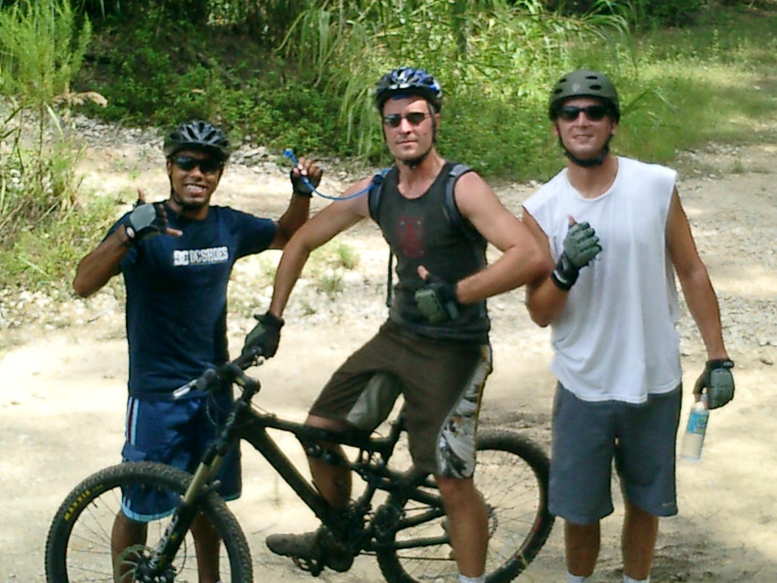 Jamis Dakar XLT: Three men stand together on a mountain biking trail, smiling and posing for the camera. Each cyclist is wearing helmets and gloves, and they are giving a thumbs up. Two of the men are on bikes, while one is holding a water bottle. The surrounding environment includes greenery and a dirt path.