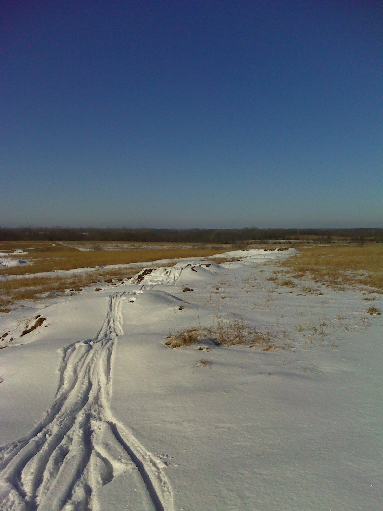 A snow-covered landscape under a clear blue sky, showing a path marked by ski or sled tracks leading through the snowy terrain, with patches of grass visible in the background. Camp Alexander Trails mountain bike trail.